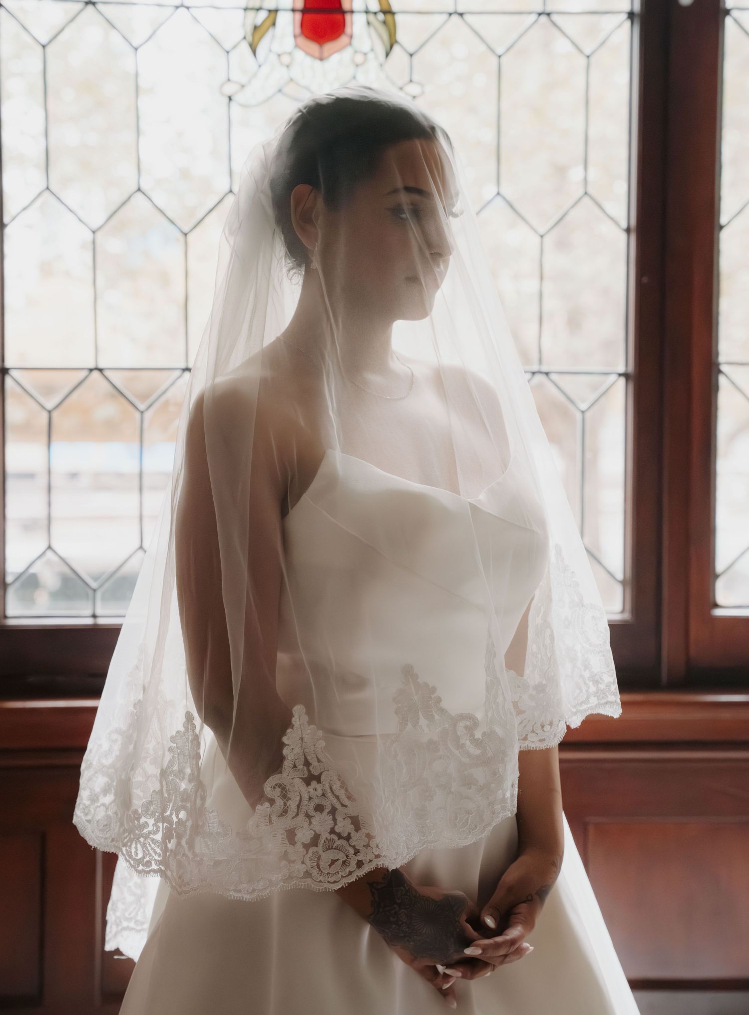 Bride wearing a white lace-trimmed veil and strapless wedding gown stands before a stained glass church window.