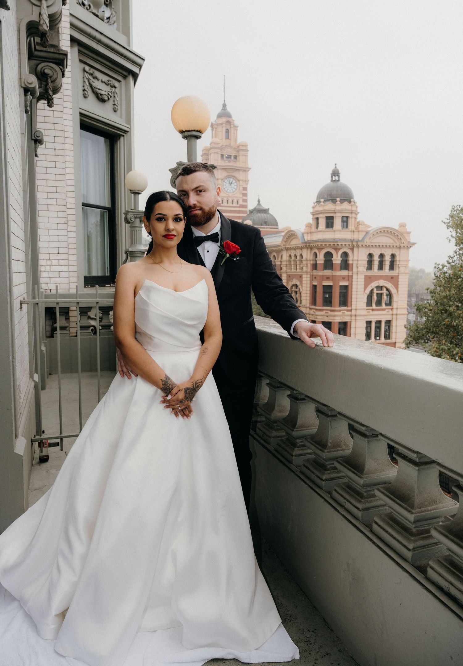 Bride in strapless white gown and groom in navy tuxedo pose on balcony with Flinders Street Station backdrop.