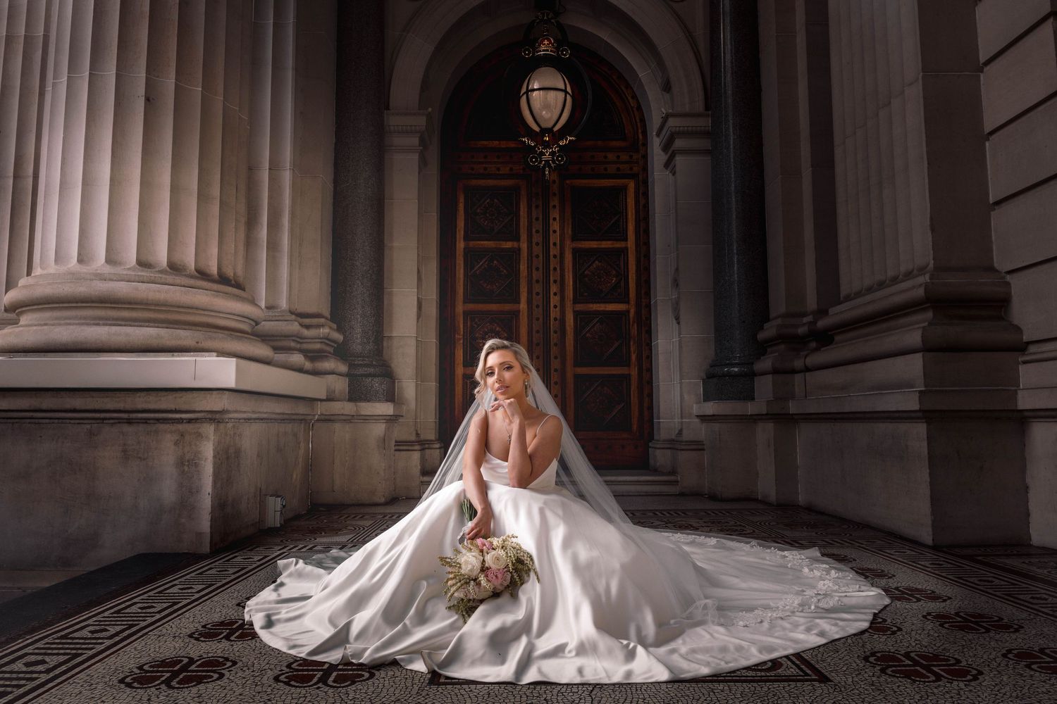 A bride in a flowing white gown with cathedral veil sits elegantly on steps of a grand historic building entrance.
