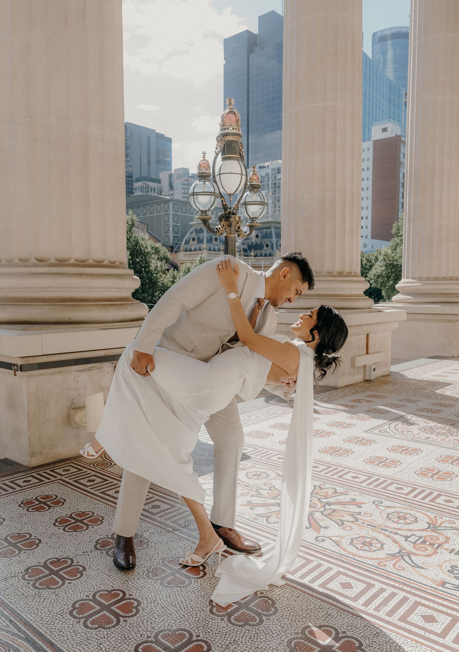 A couple in elegant white attire dances gracefully on an ornate tiled terrace with city skyscrapers in the background.