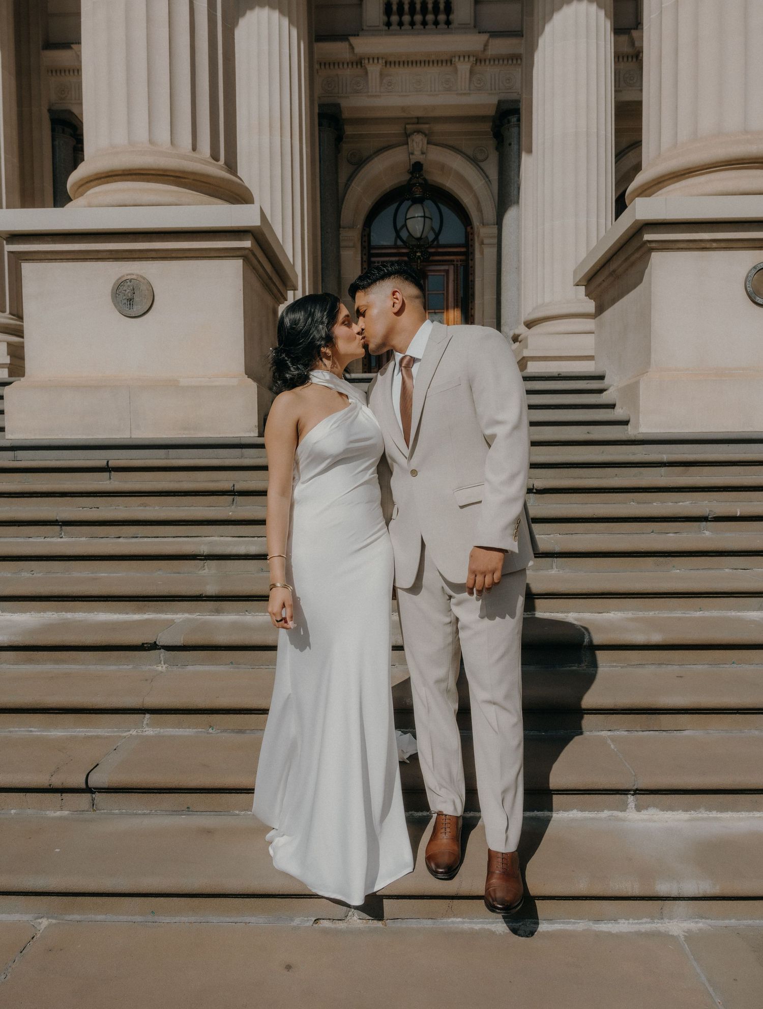 A couple in white attire shares a romantic moment on elegant stone steps of a grand building.
