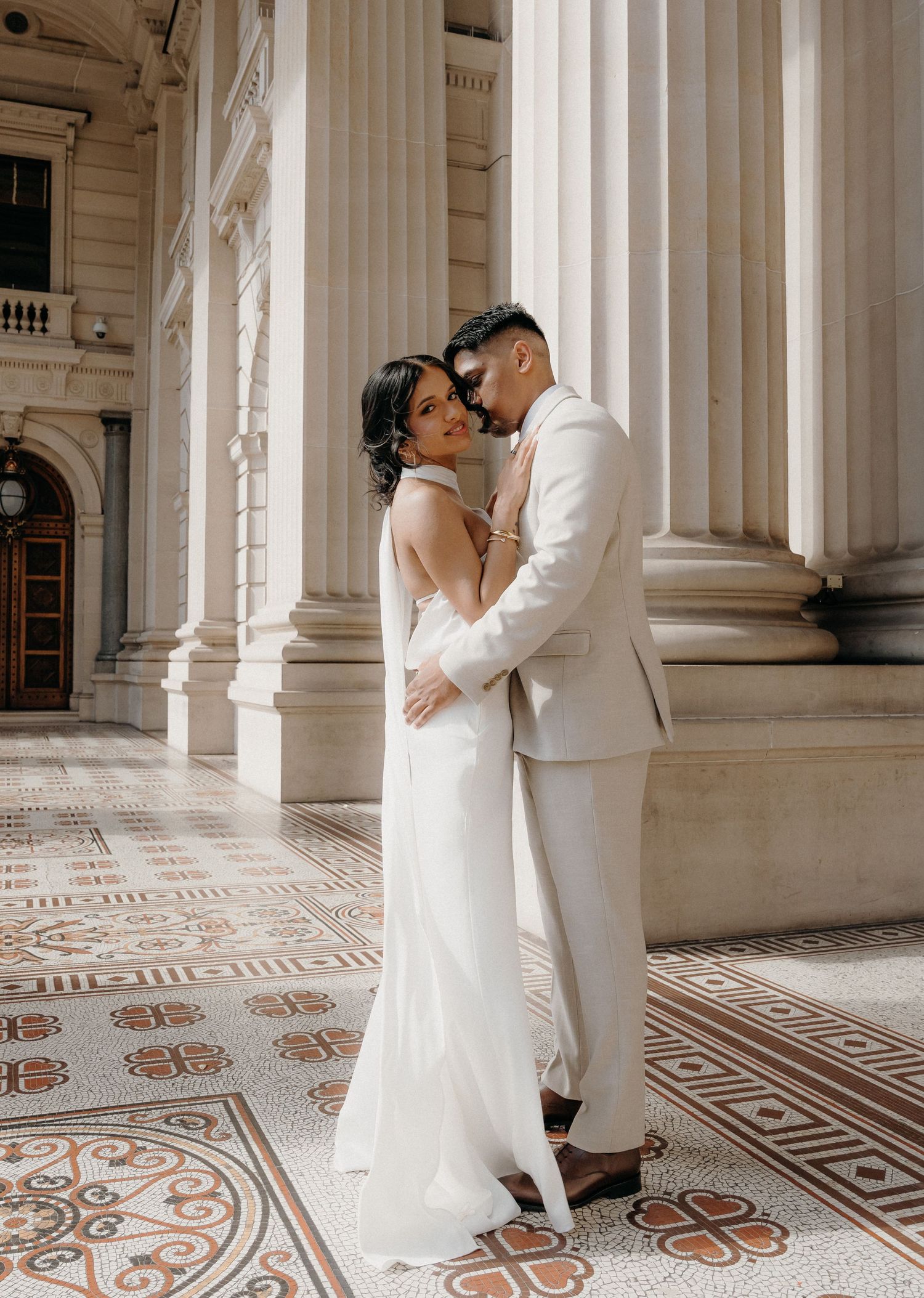 A romantic wedding photo at a grand building with ornate columns and decorative tiled flooring.