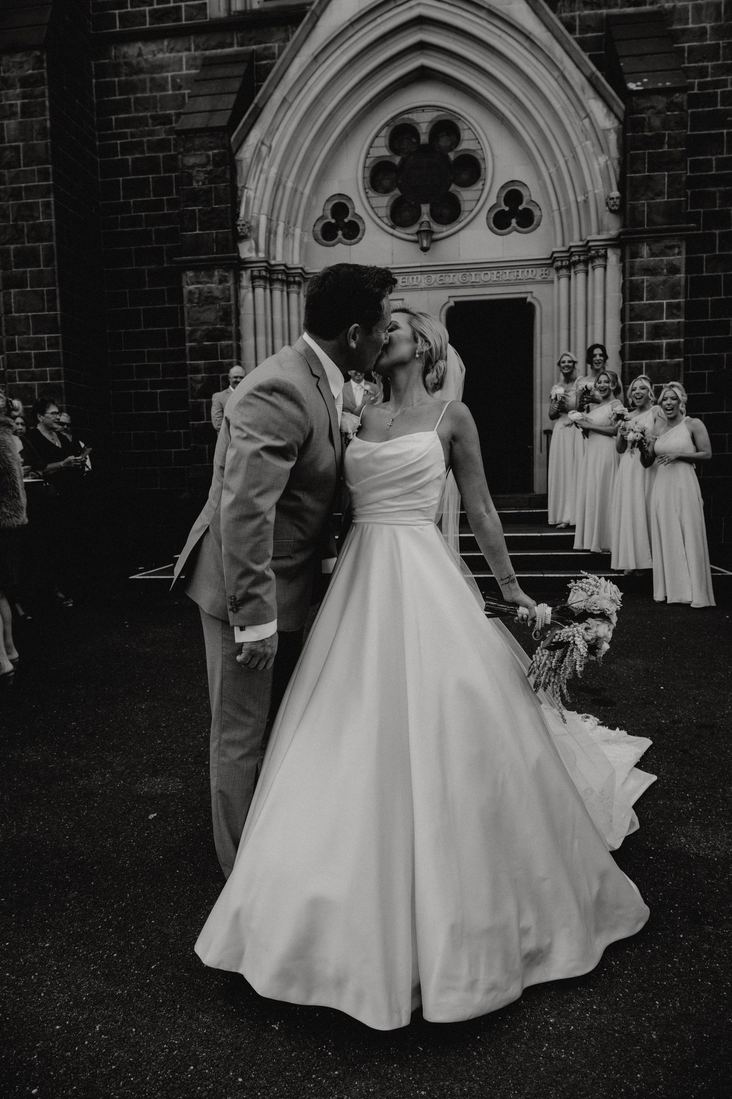 A romantic black and white wedding photo of a couple sharing a kiss outside a gothic church with bridesmaids in background.
