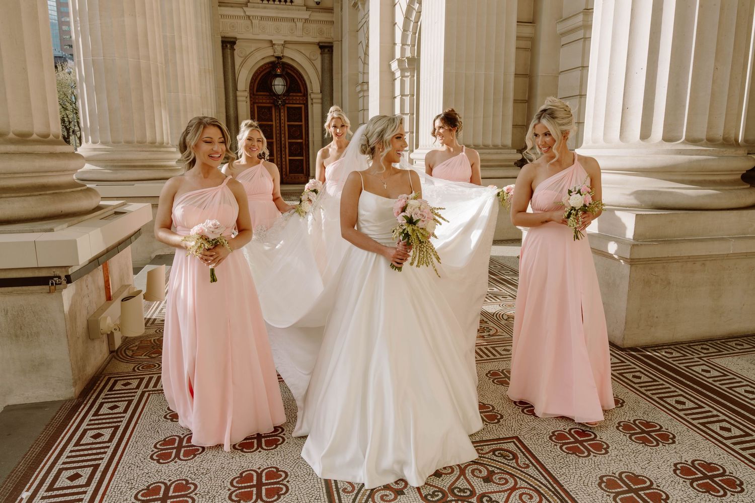 A wedding party in pink bridesmaid dresses and white bridal gown pose together in an elegant marble hallway.