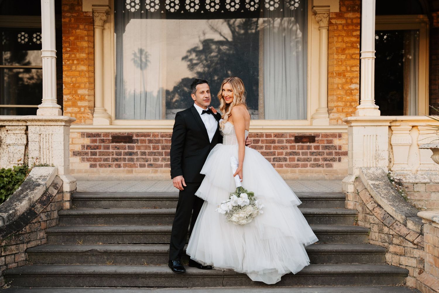 A newlywed couple shares a romantic moment on the steps of a brick building with white columns and elegant windows.