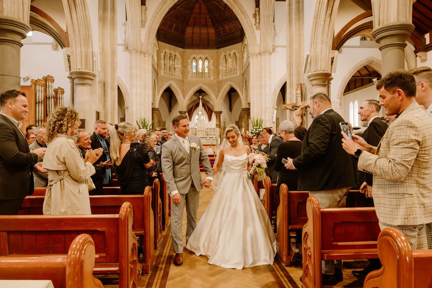 A traditional wedding ceremony takes place in a grand cathedral with red pews and vaulted ceilings.