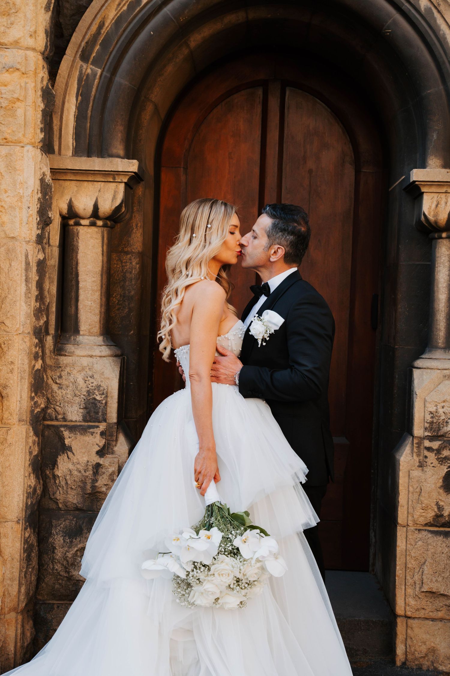 A romantic wedding moment captured in front of a historic stone church with arched doorways and ornate architecture.