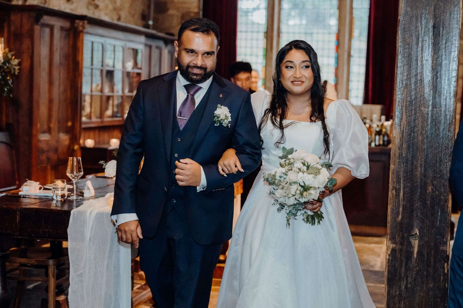 A wedding couple walks between dark wooden pillars in a rustic venue with soft ambient lighting.