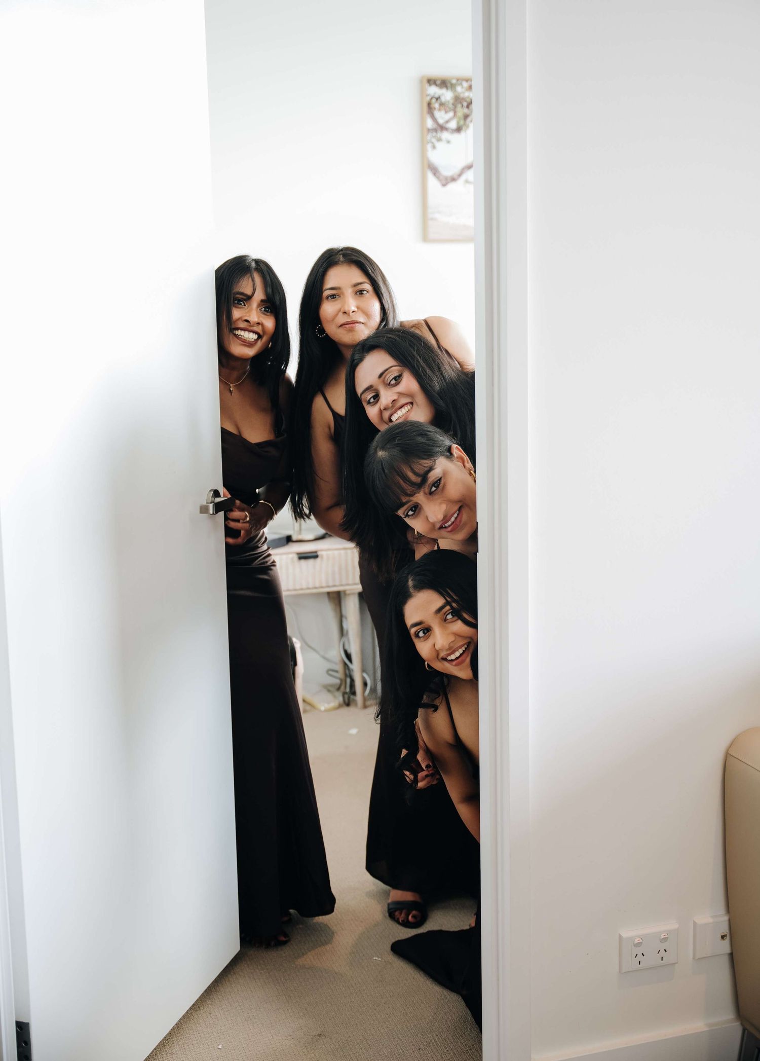 A group of friends peek playfully around a doorway corner in a bright interior space.