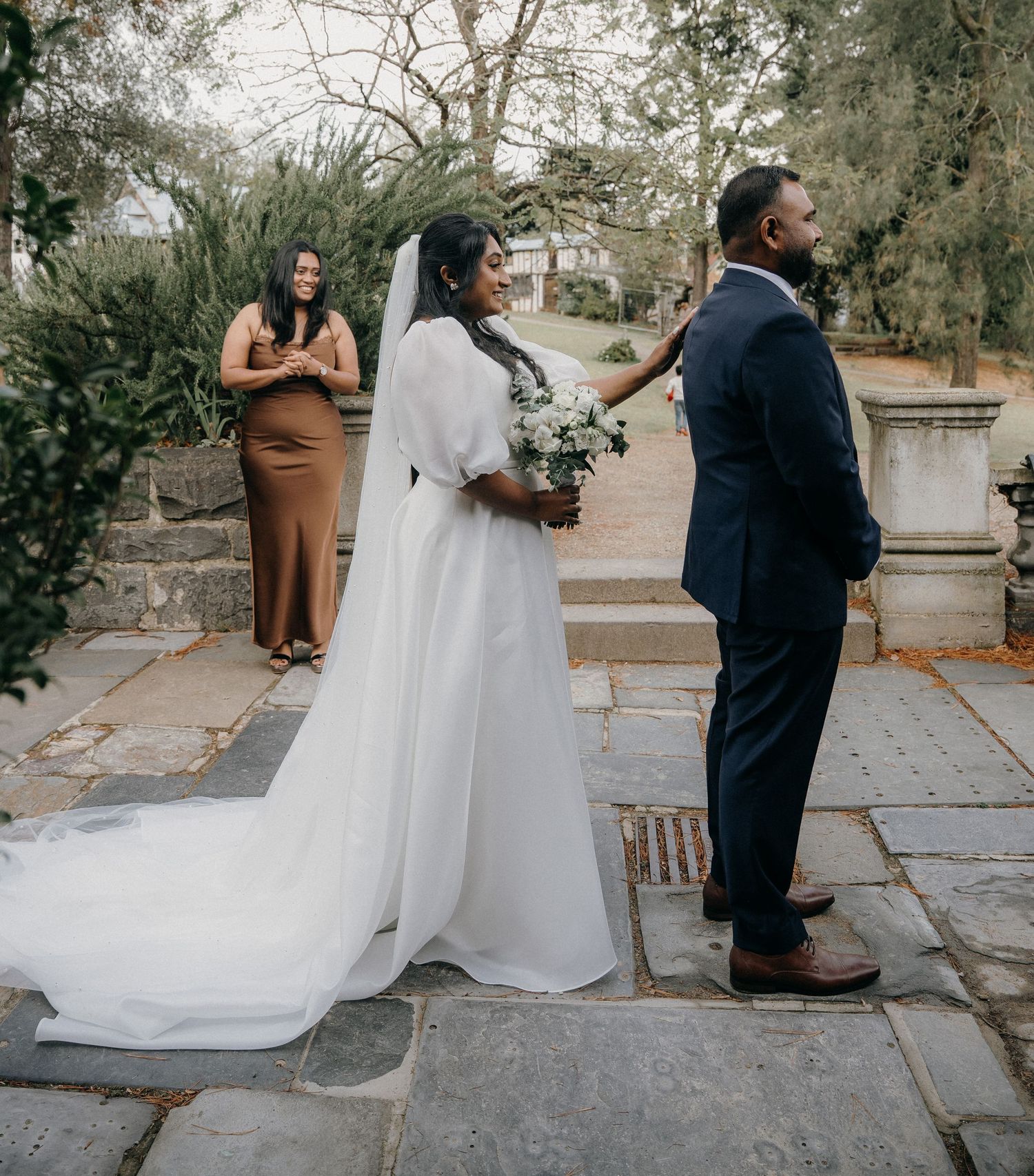 A couple in formal wedding attire poses for creative first look photos in a garden setting.