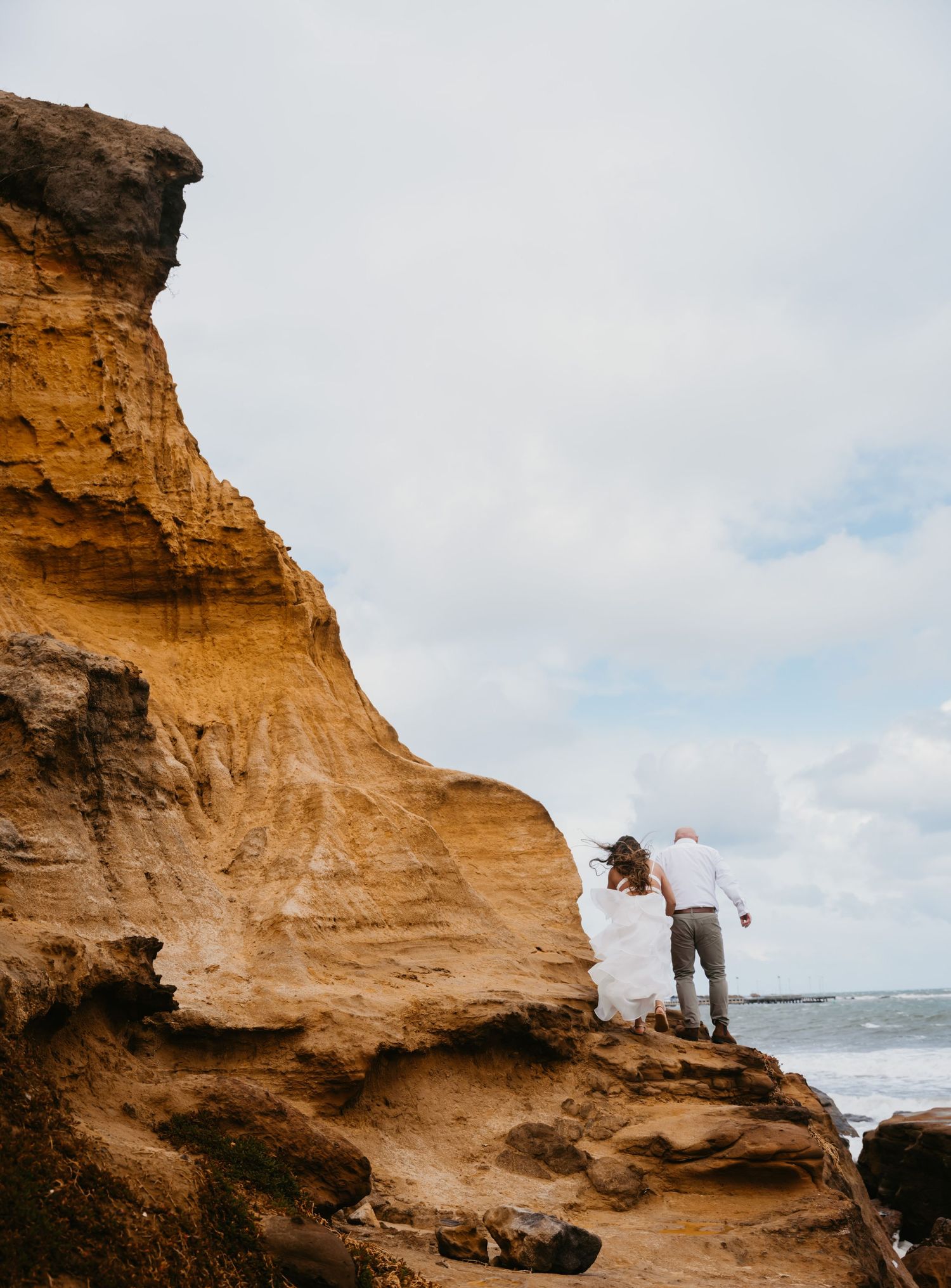 Couple standing on dramatic sandstone cliffs overlooking the ocean during a cloudy day.