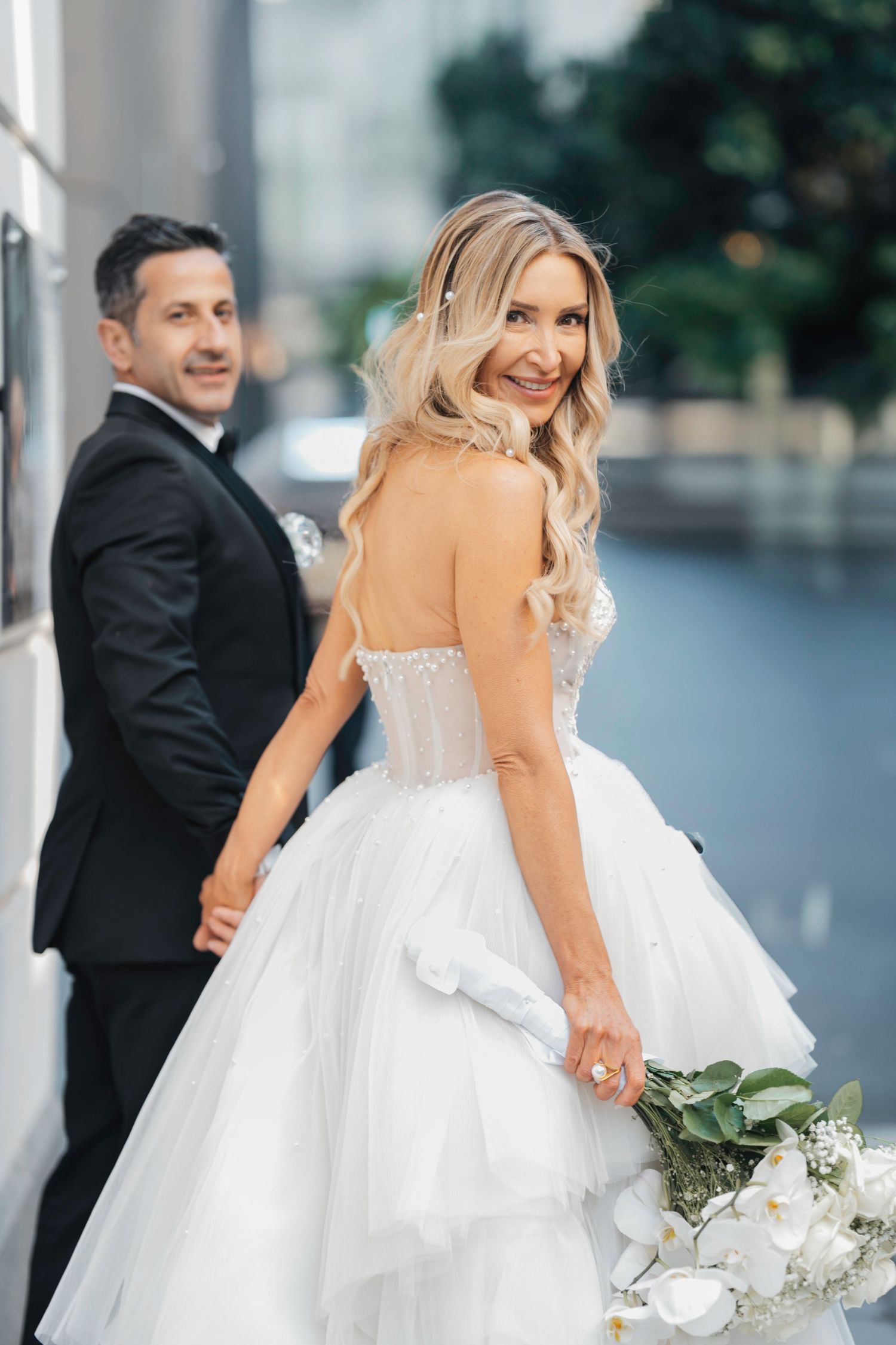 A bride in a flowing white gown poses with a bouquet while a groom in a black suit stands behind her.