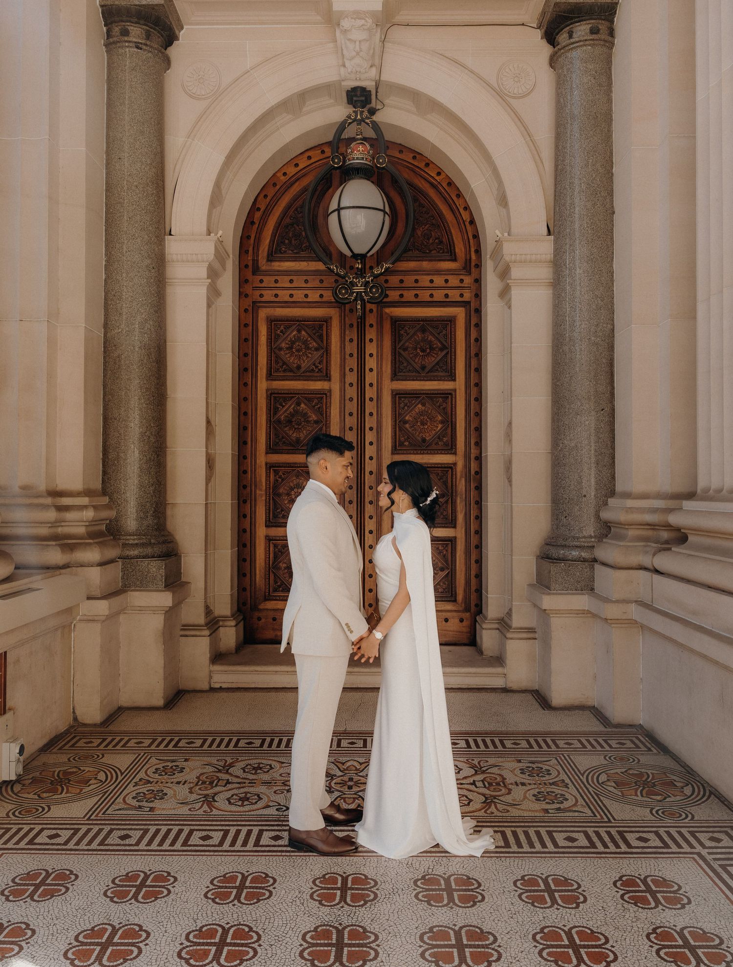 Two figures in white robes stand in an elegant hallway with ornate marble floors and arched wooden doors.