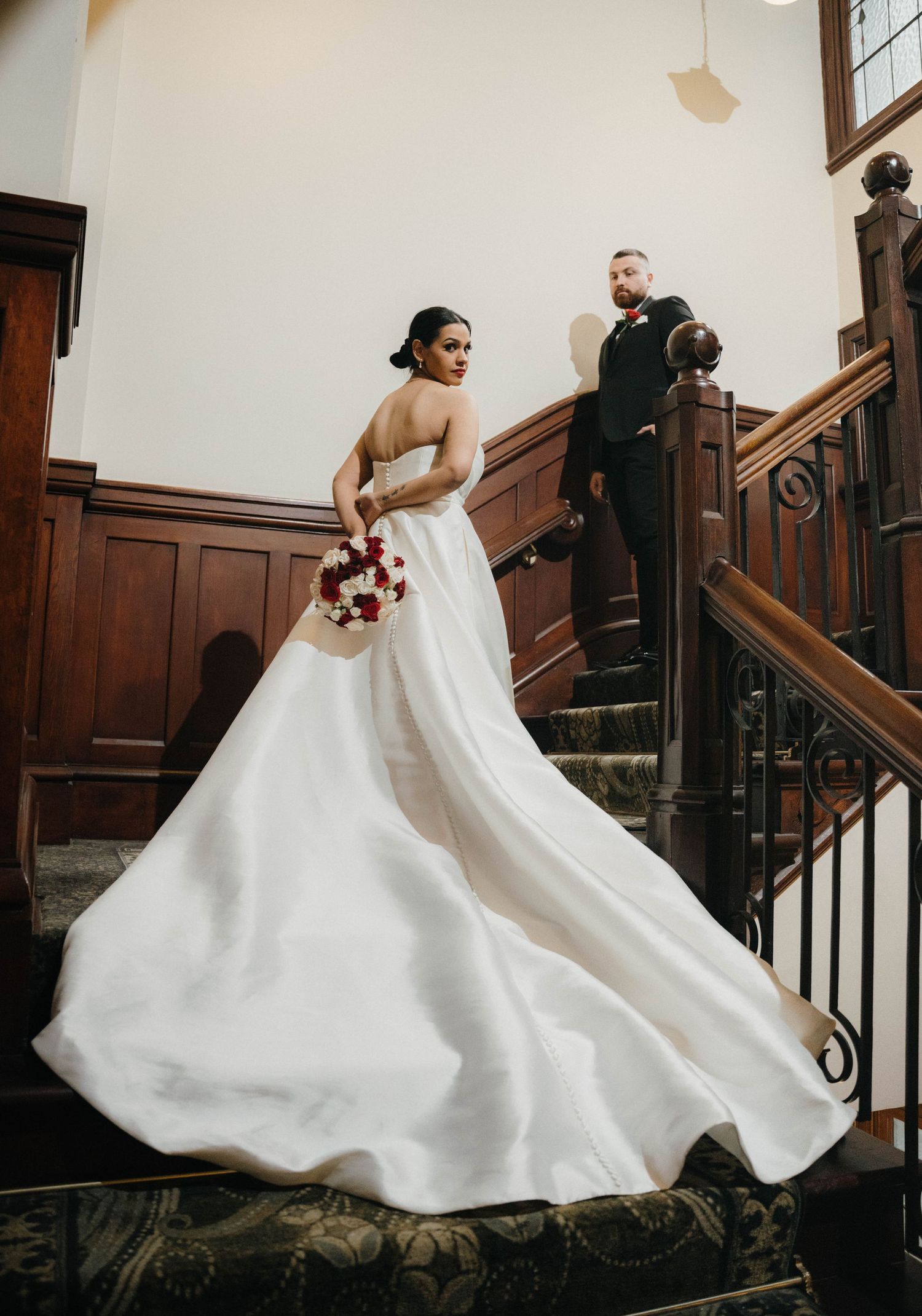 Bride in elegant white strapless ball gown holding red bouquet on staircase, groom in black suit standing behind her.