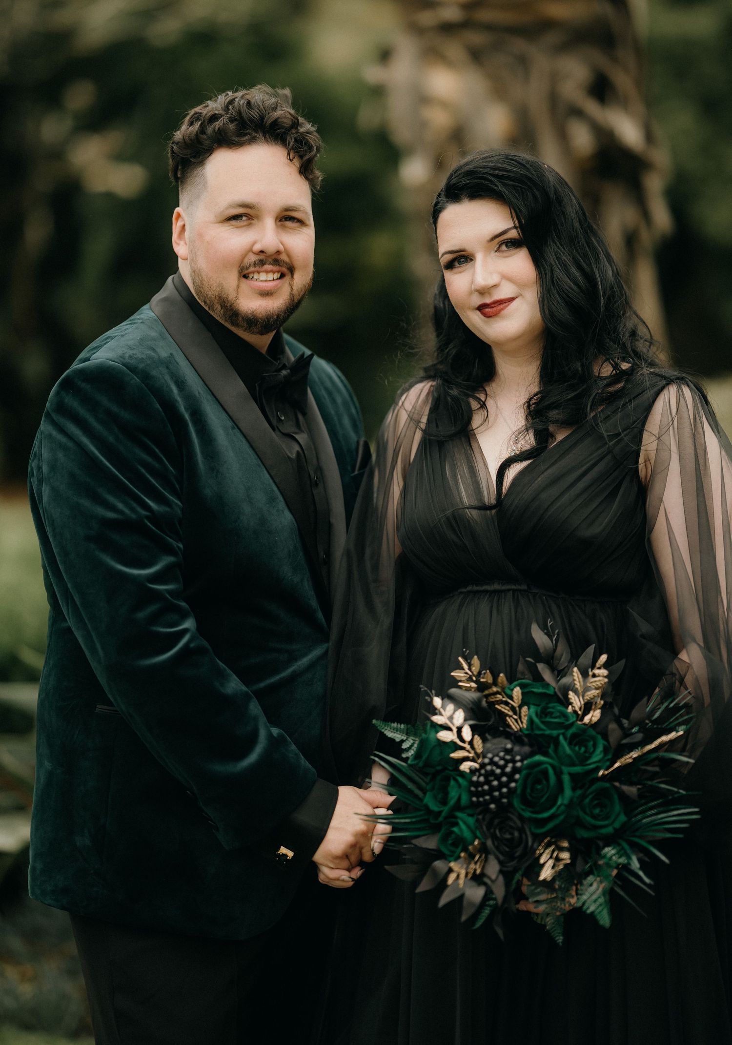 A couple in dark attire poses together in a romantic outdoor setting with moody fall-inspired florals.