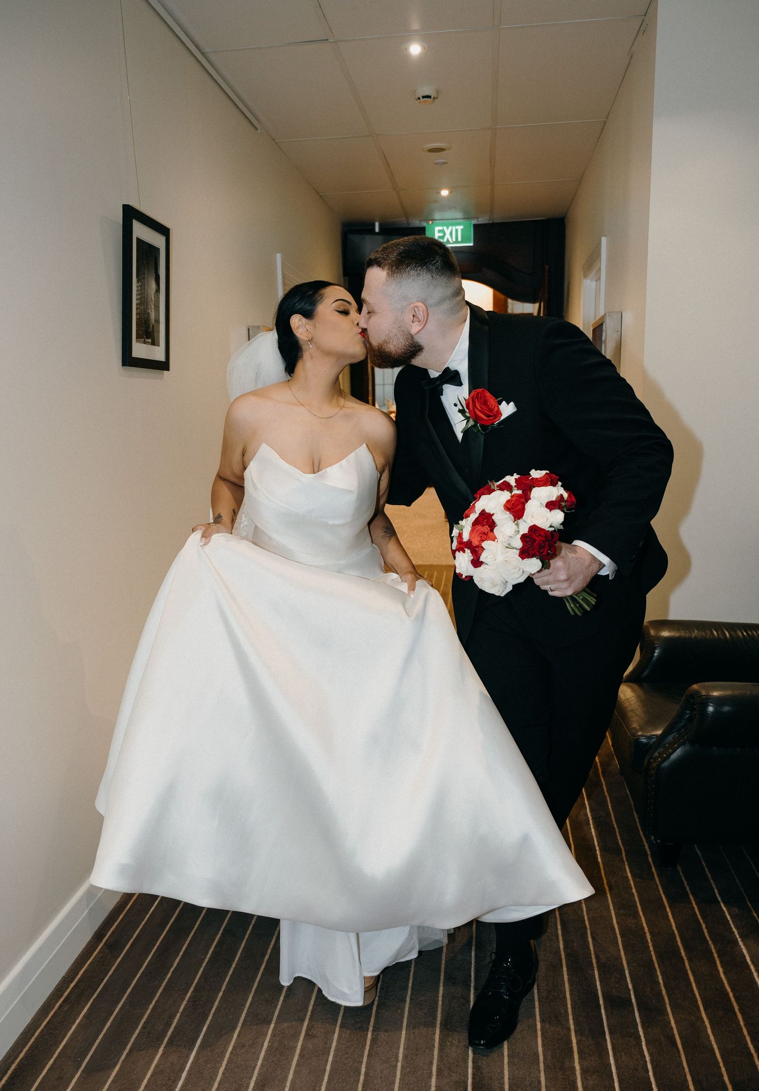 Bride and groom sharing a kiss in a hotel hallway, bride in strapless white gown, groom in black tux holding red bouquet.