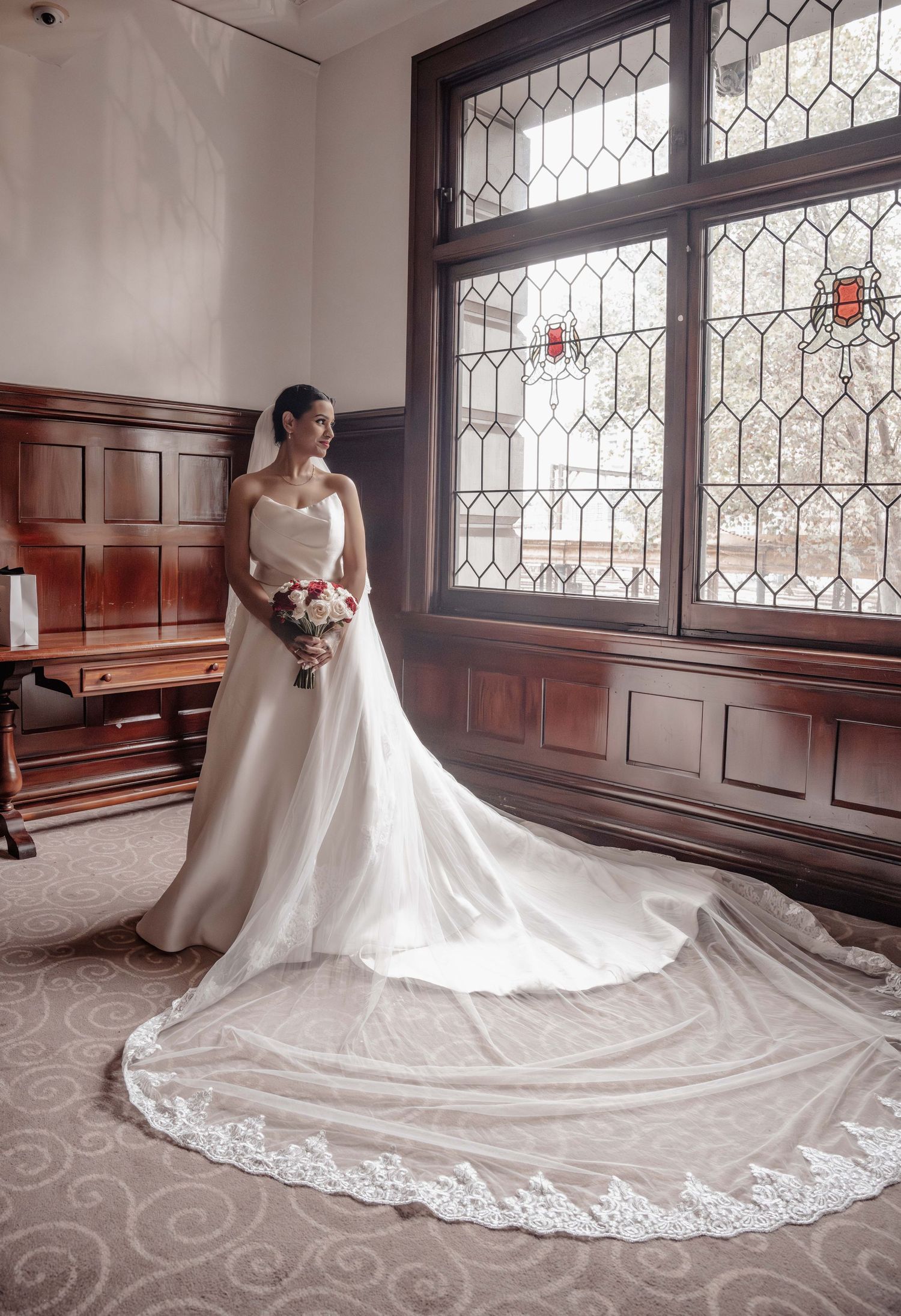 Bride in elegant strapless white gown with long lace-trimmed cathedral veil holding bouquet by leaded glass windows.