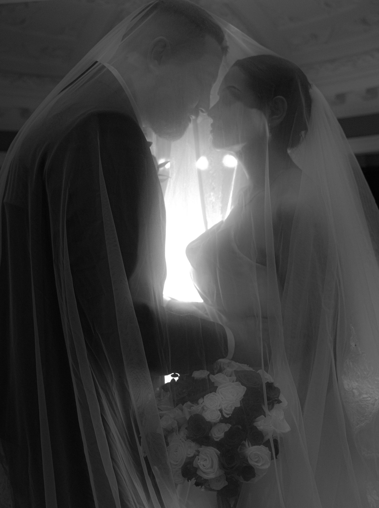 Black and white wedding photo of bride and groom sharing intimate moment under veil, bride holding rose bouquet.