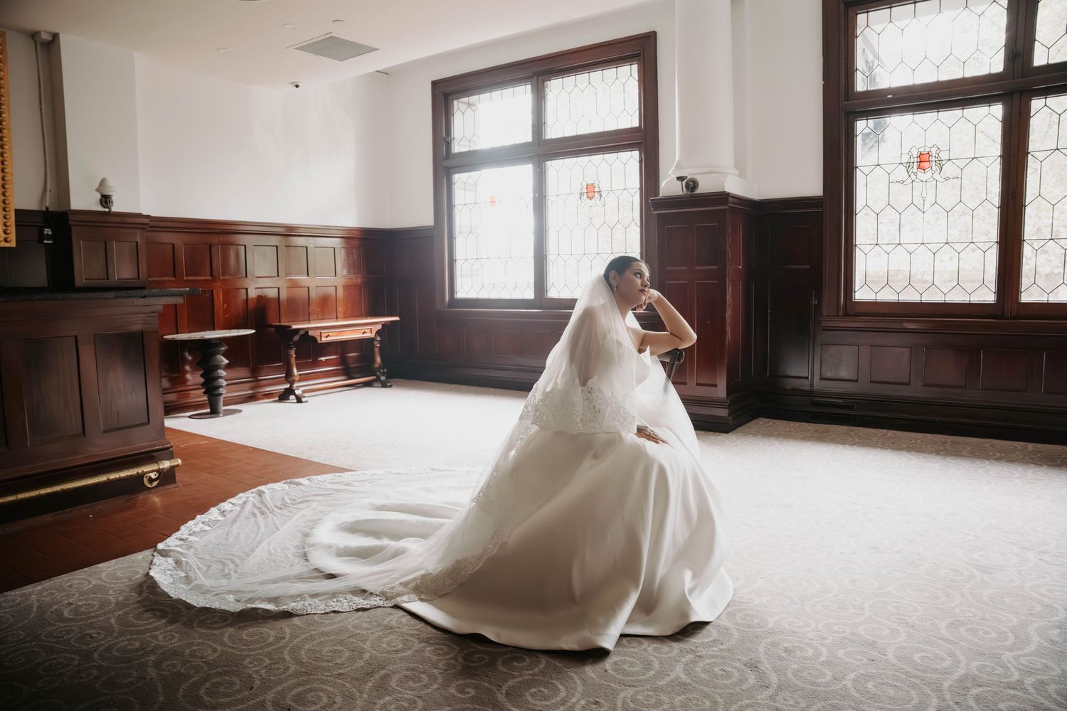 Bride in elegant white wedding gown with long veil posing in a grand room with stained glass windows.