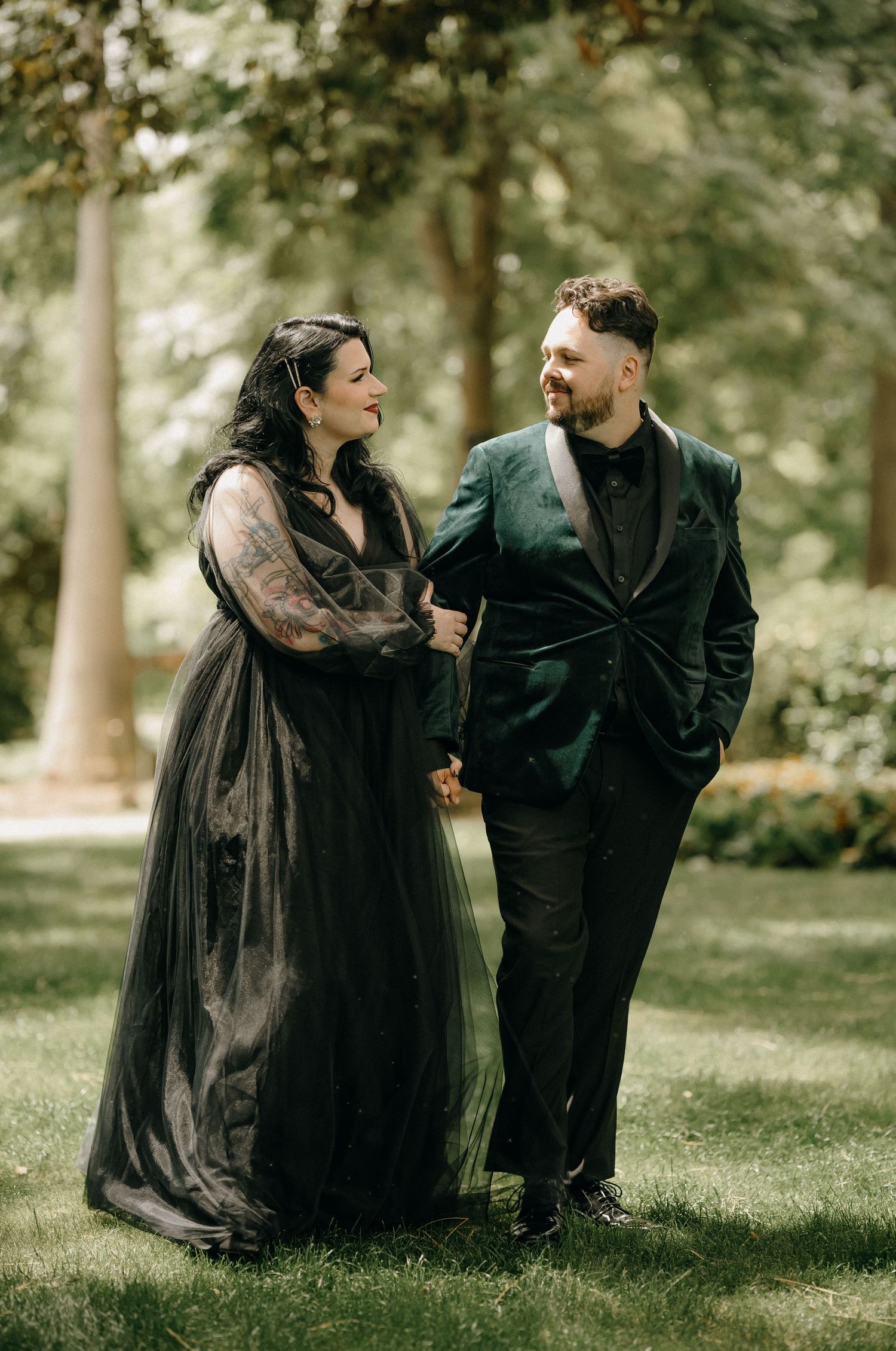 A couple in gothic-inspired black attire shares intimate moments on a grassy lawn in a wooded setting.