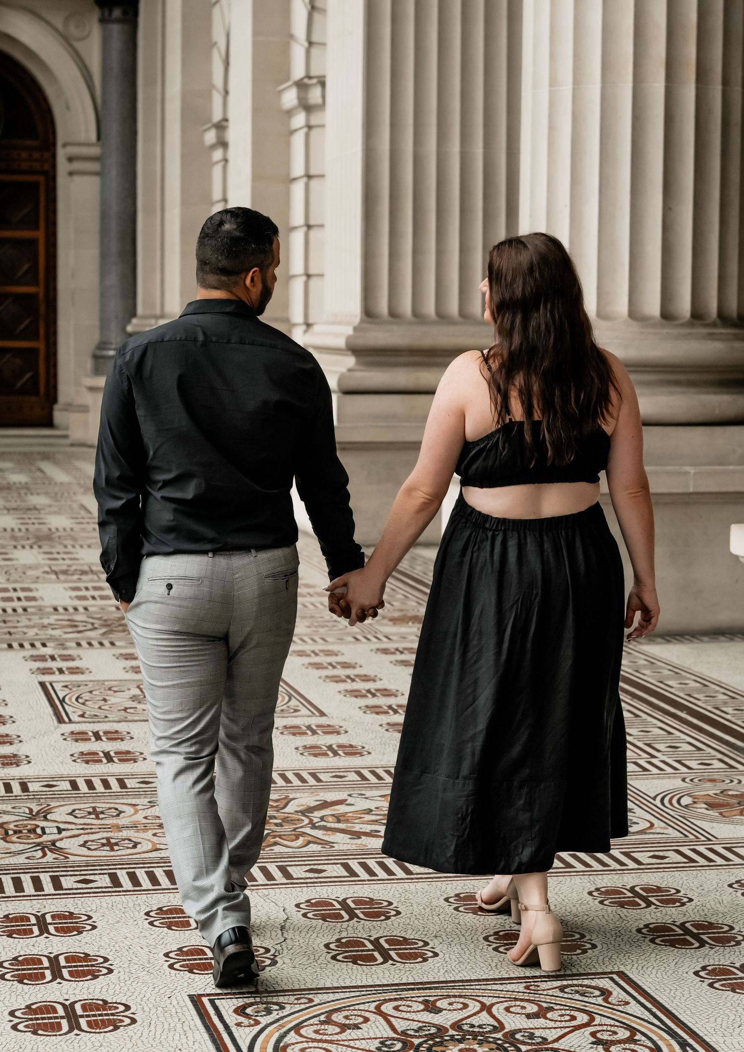 Two people in formal attire walk together on an ornate patterned floor in a historic building with white columns.