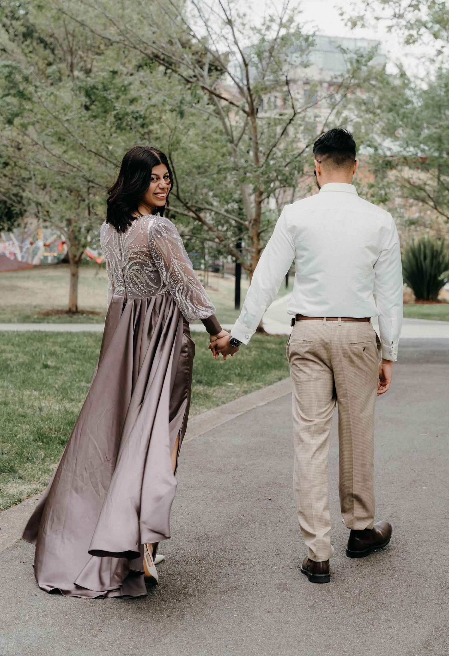 A couple holding hands and walking on a path, with the woman wearing a flowing mauve dress and man in white shirt.