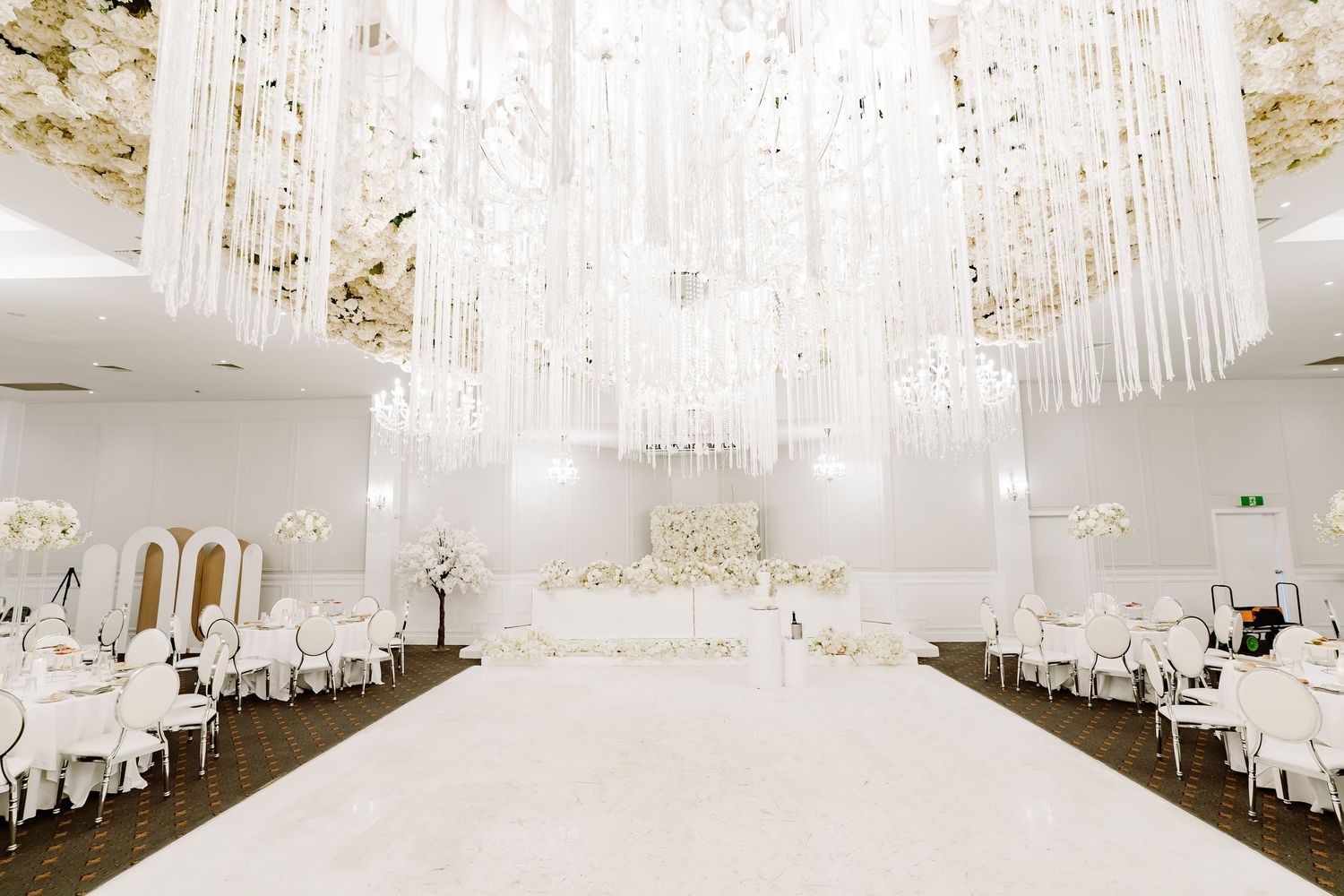 Elegant white wedding reception venue with crystal chandeliers and white floral centerpieces on round tables.