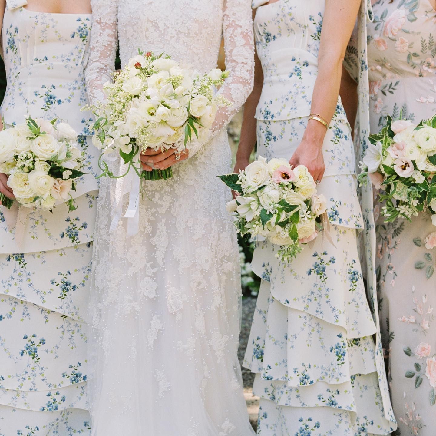 Row of bridesmaids in light blue floral dresses holding white and blush bouquets at outdoor wedding ceremony.