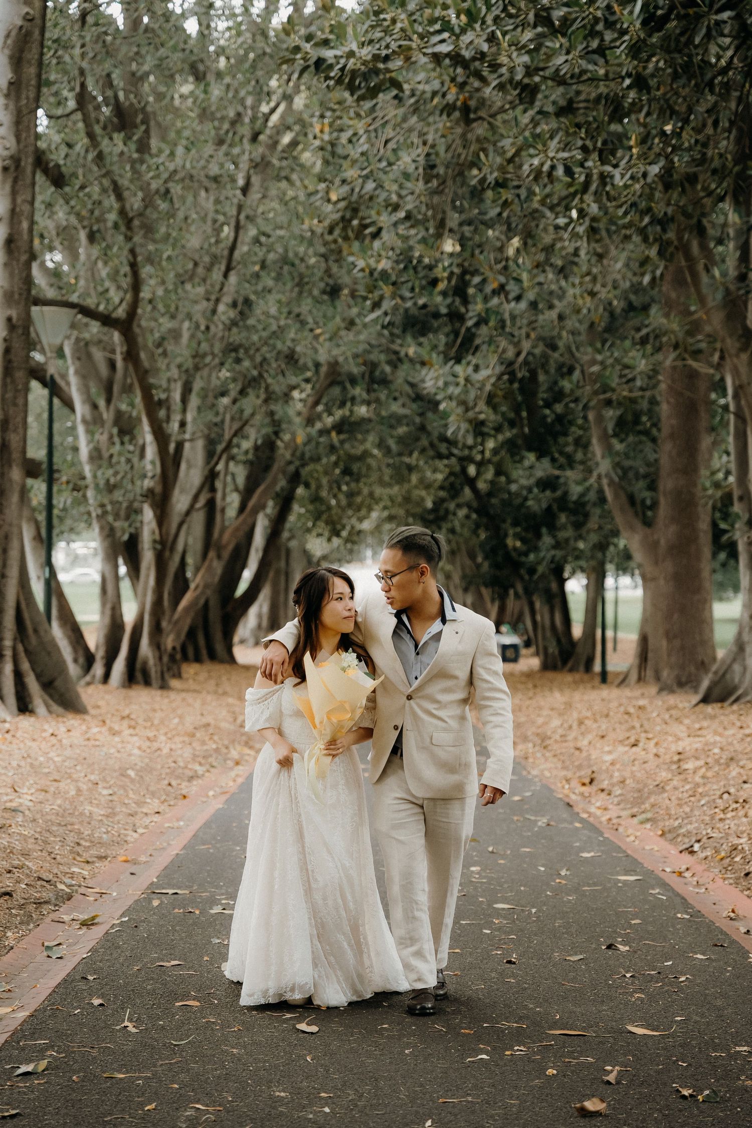 A couple in white attire shares romantic moments while walking down a tree-lined path during a photo session.