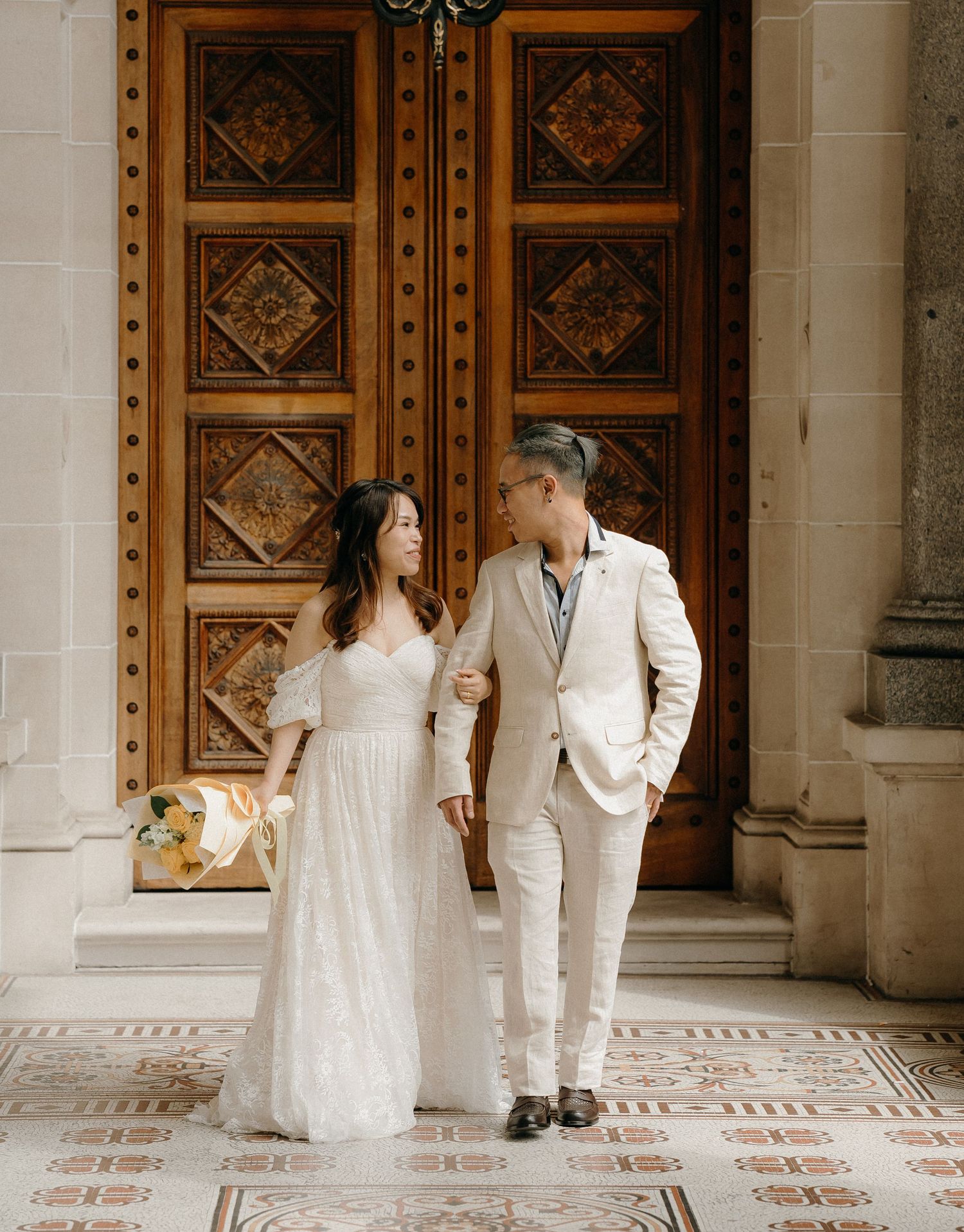 A couple in wedding attire stands together on an ornate tiled floor in front of decorative wooden doors.
