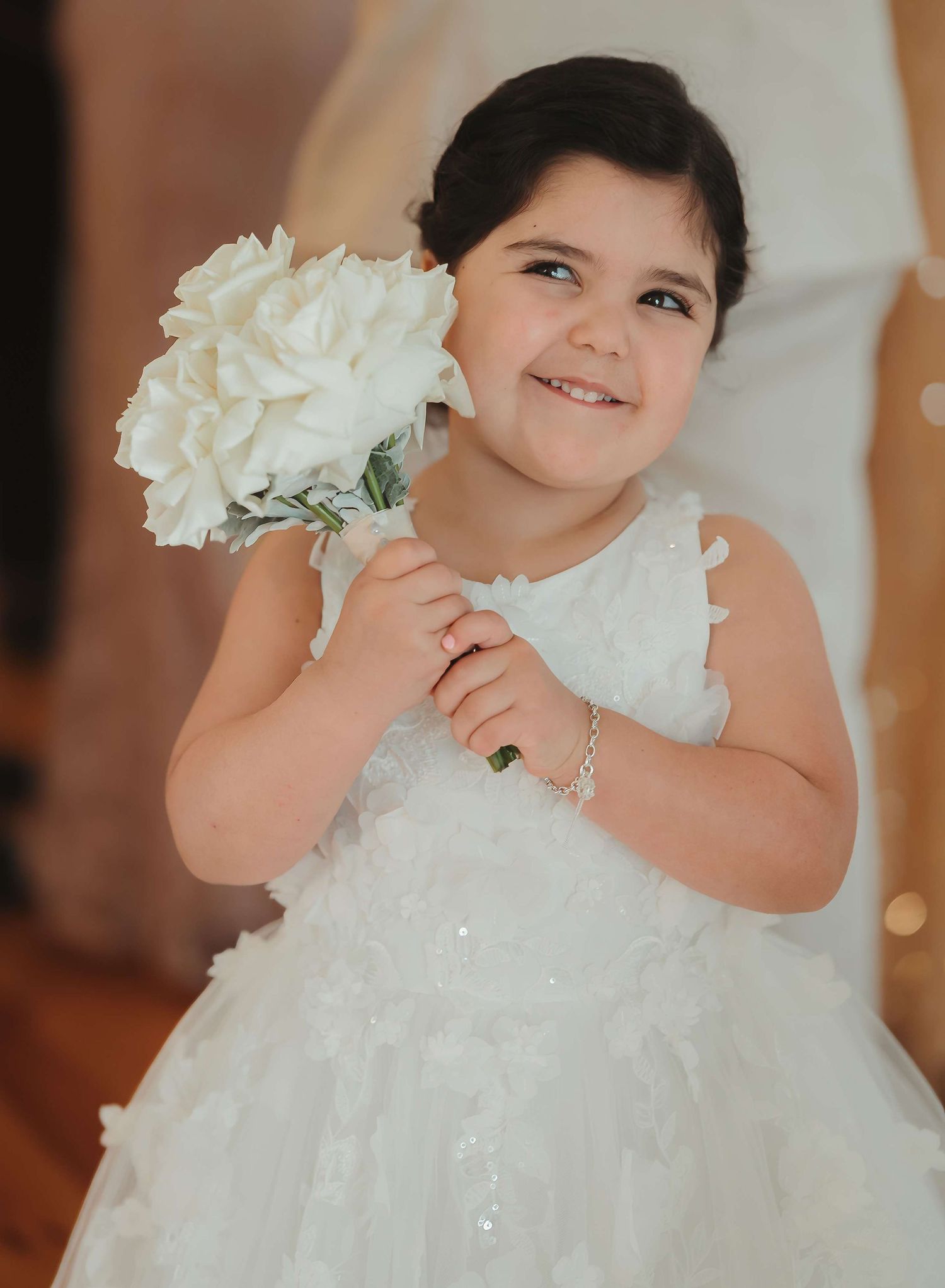 A child wearing a sparkly white dress holds a white hydrangea bouquet while smiling.