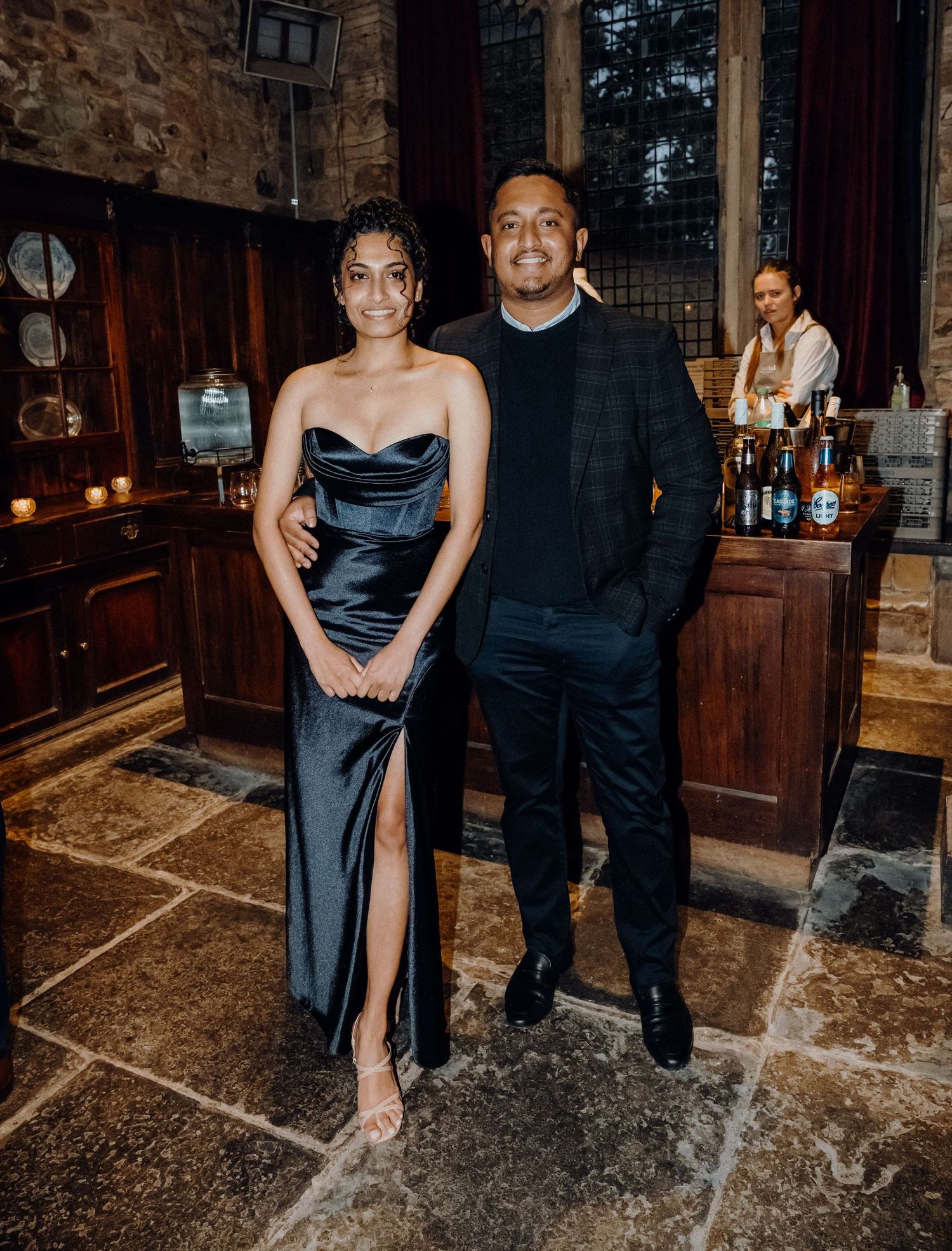 A couple pose together at an elegant event, with the woman wearing a navy satin gown and the man in all black attire.
