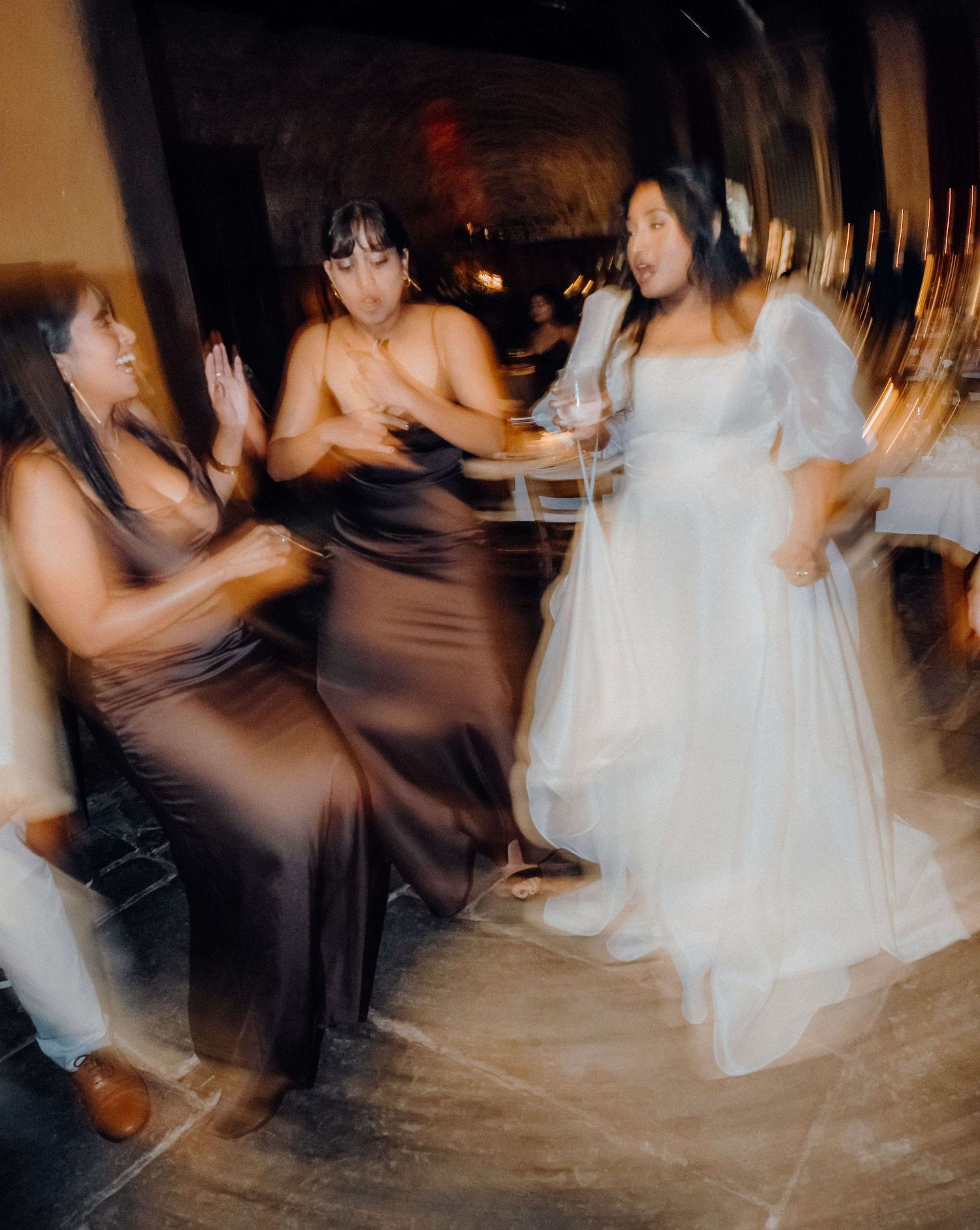 Motion-blurred photograph of people dancing energetically at a wedding reception, with one in a white dress and others in dark gowns.