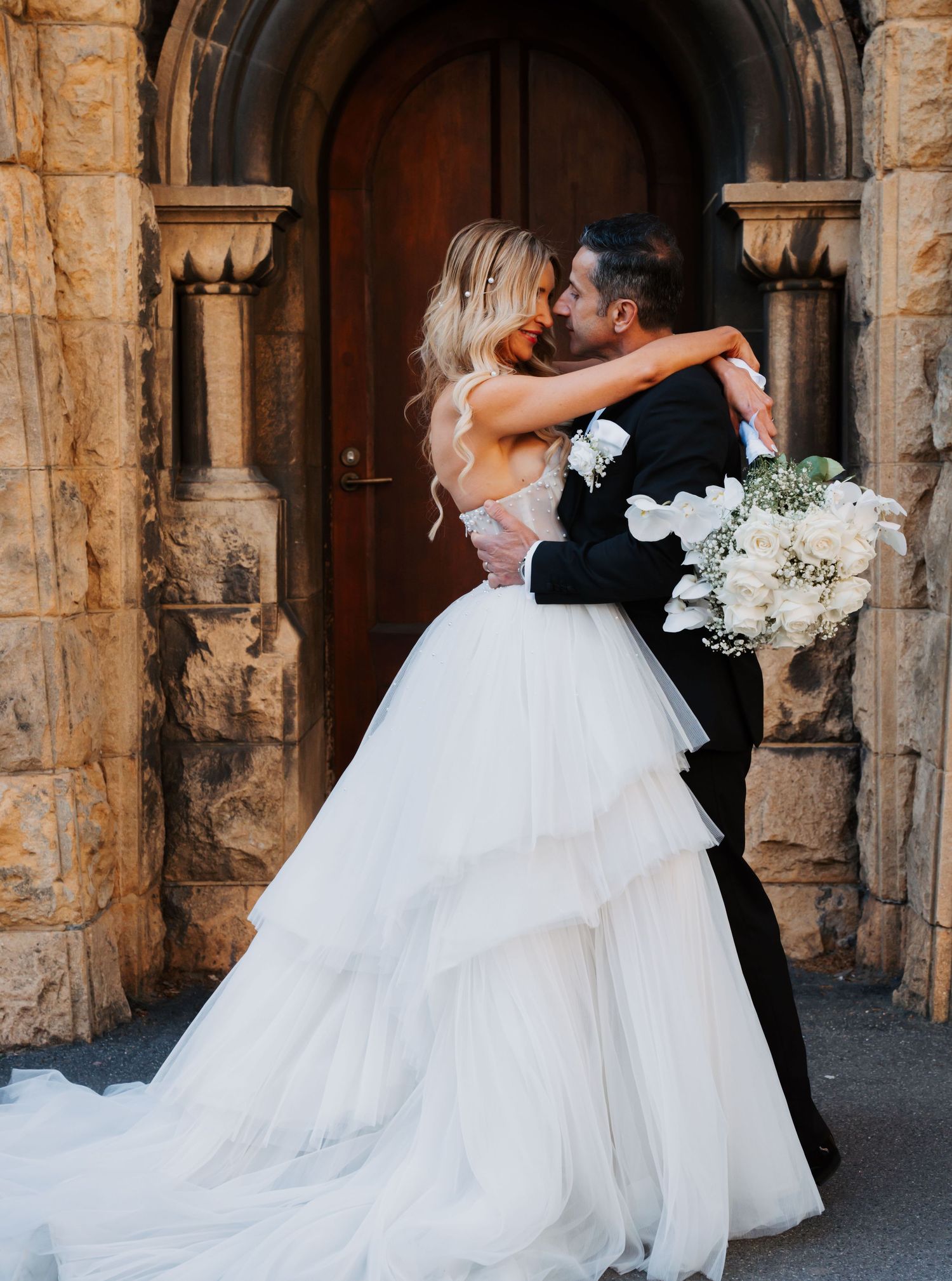 A romantic wedding couple embraces in front of an ornate stone archway, with the bride wearing a flowing white ballgown.