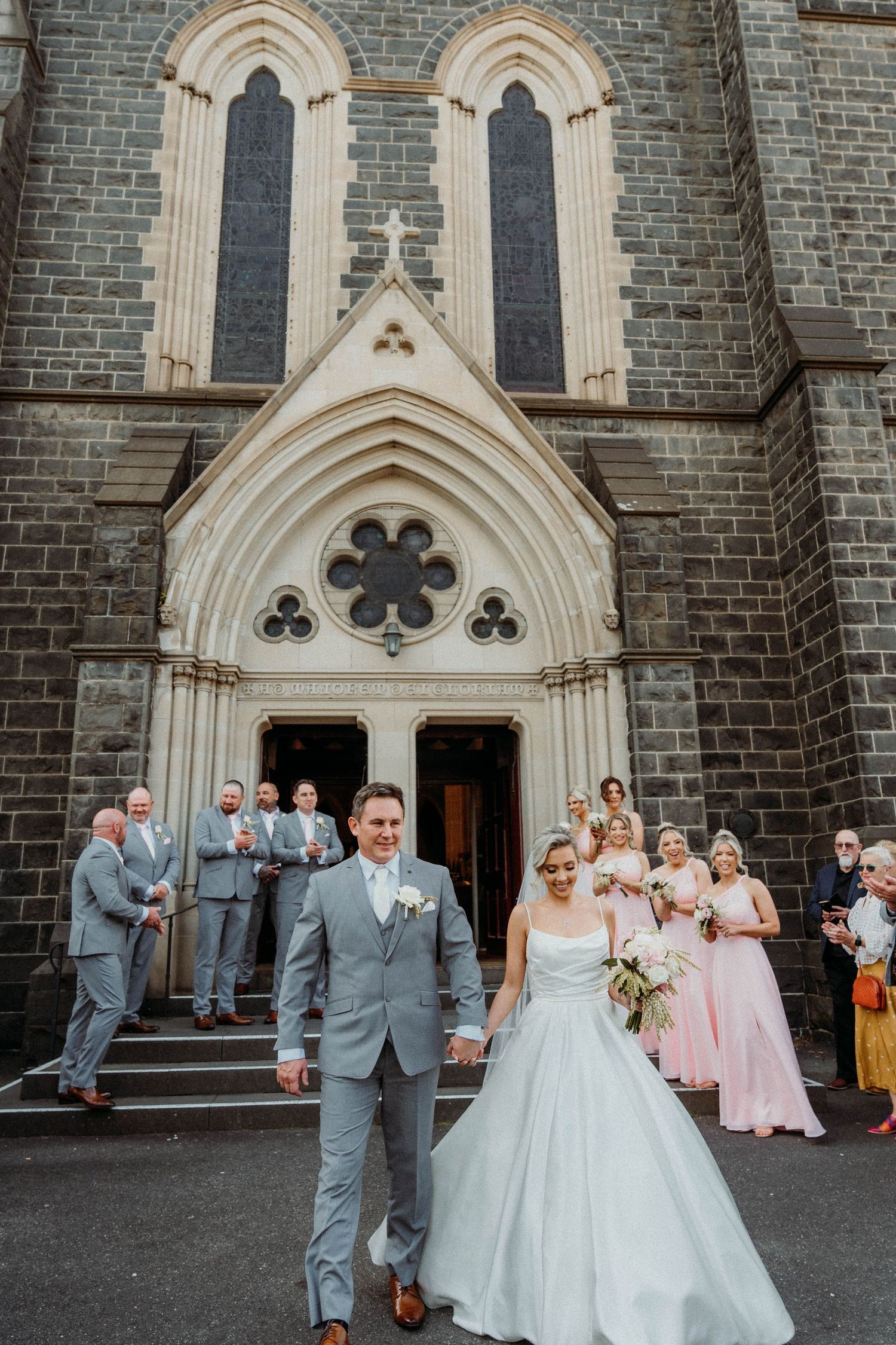 Wedding party gathered outside a Gothic stone church with arched doorways and windows in a traditional ceremony setting.