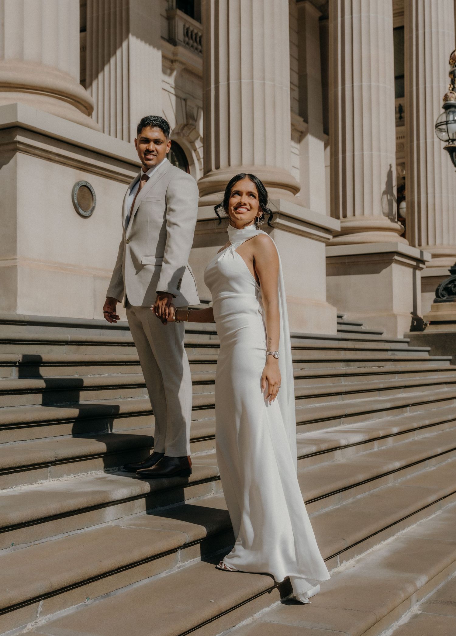 Elegant couple in white wedding attire pose on grand marble steps of a historic building with classical columns.