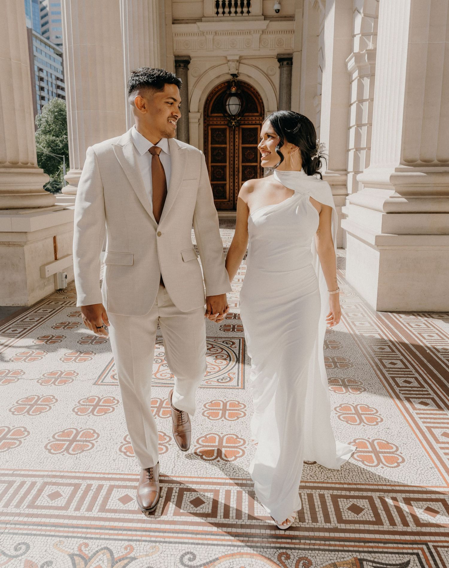 A couple in matching white wedding attire walks through an ornate corridor with patterned tile floors and columns.
