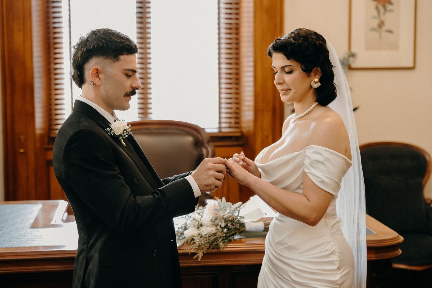 Groom placing wedding ring on brides finger during intimate courthouse ceremony, bride in elegant off-shoulder white gown.