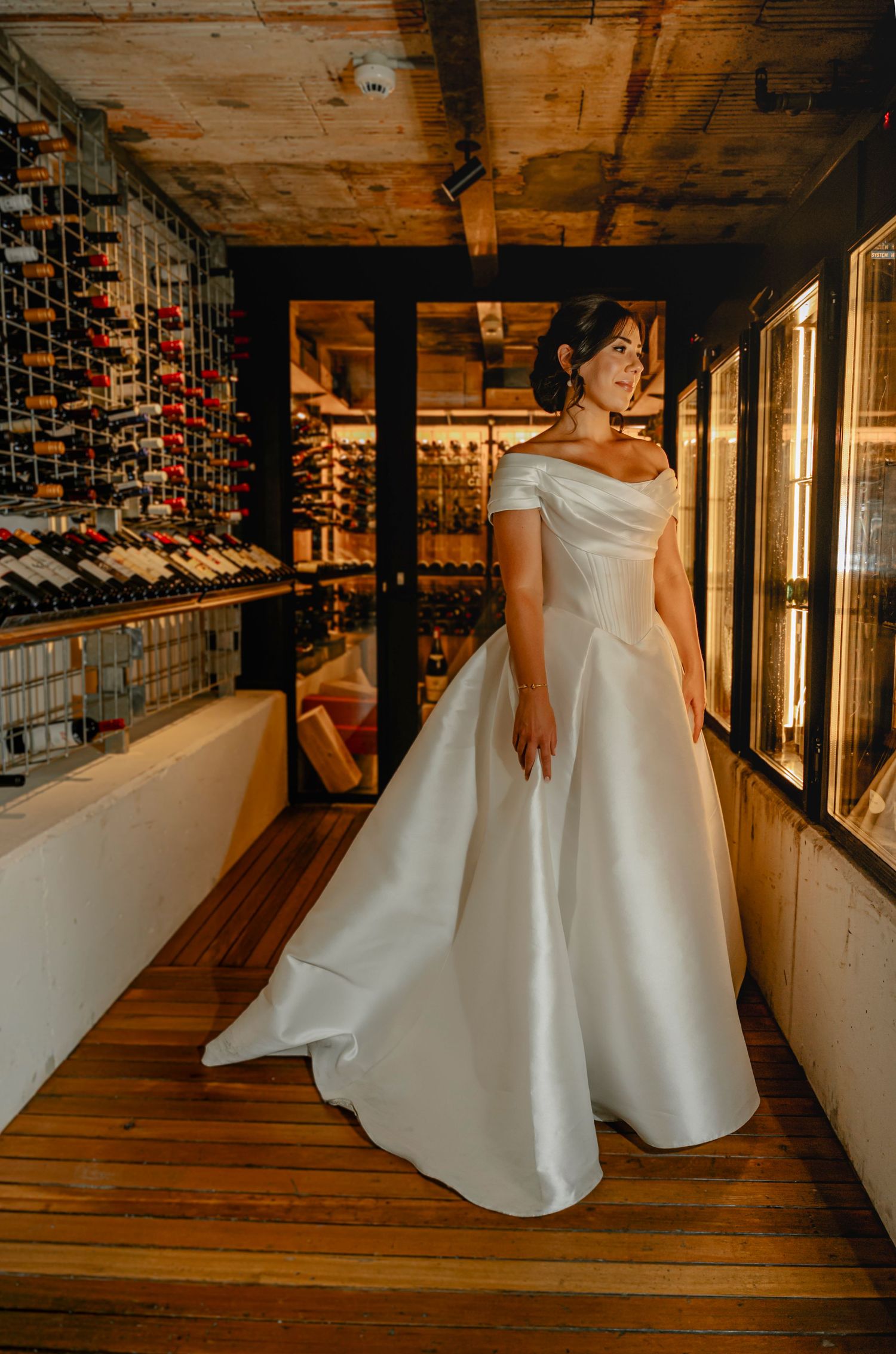 Someone in a white wedding dress poses elegantly in a rustic venue with exposed wooden beams and wine storage.