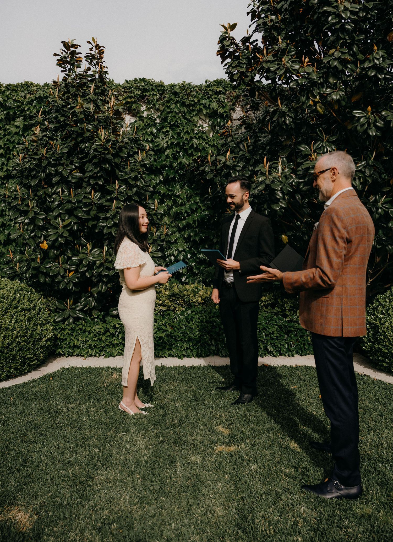 A couple stands together in a garden setting with lush greenery and trees in the background.