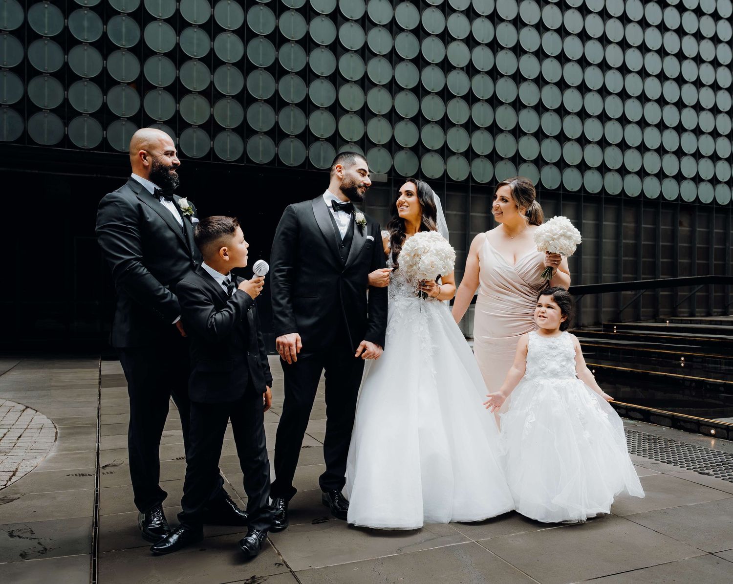 Wedding party in formal attire including white gowns and black suits sharing a fun moment against modern architectural background.
