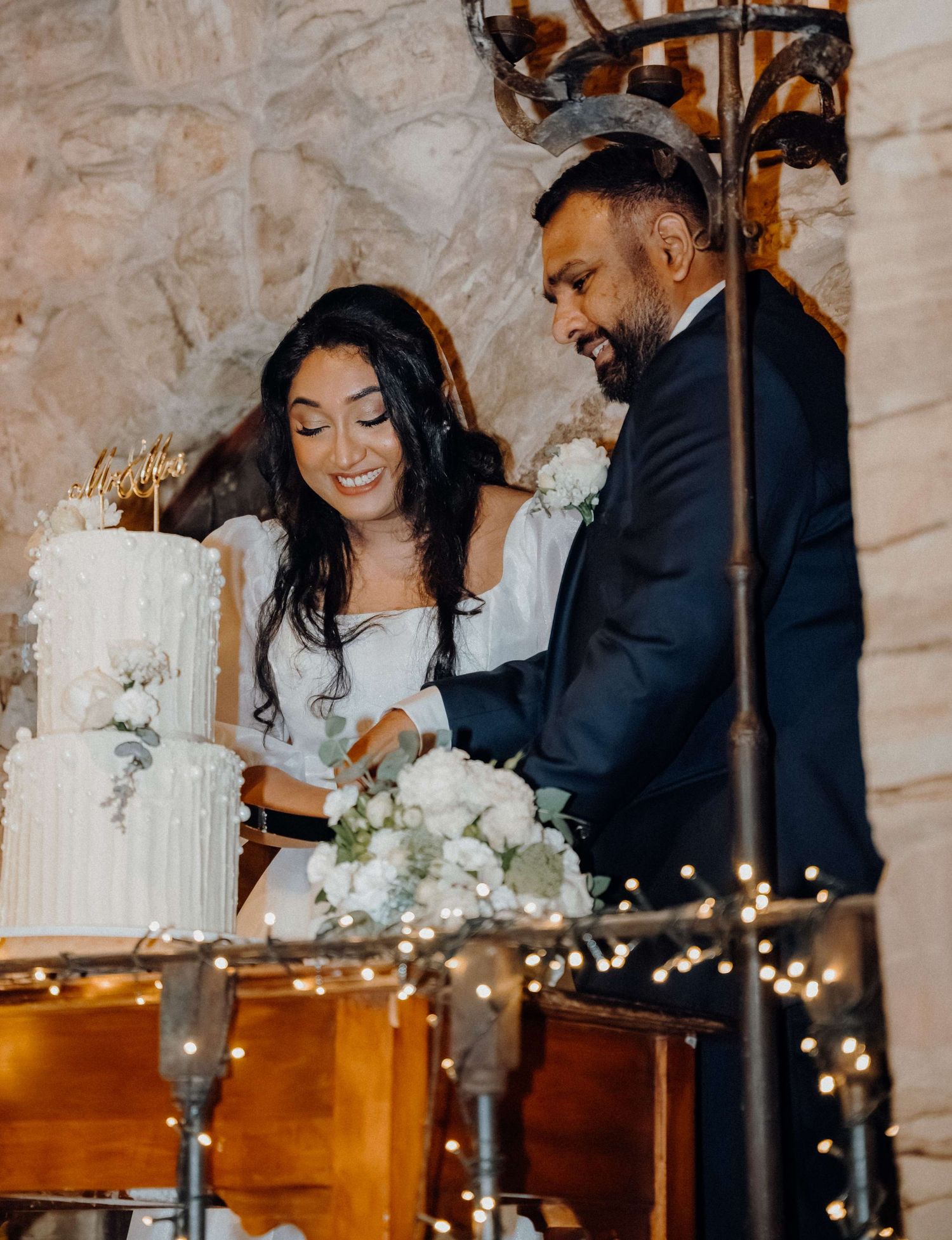 A couple cuts their elegant white wedding cake together while standing at a reception table decorated with twinkling lights.