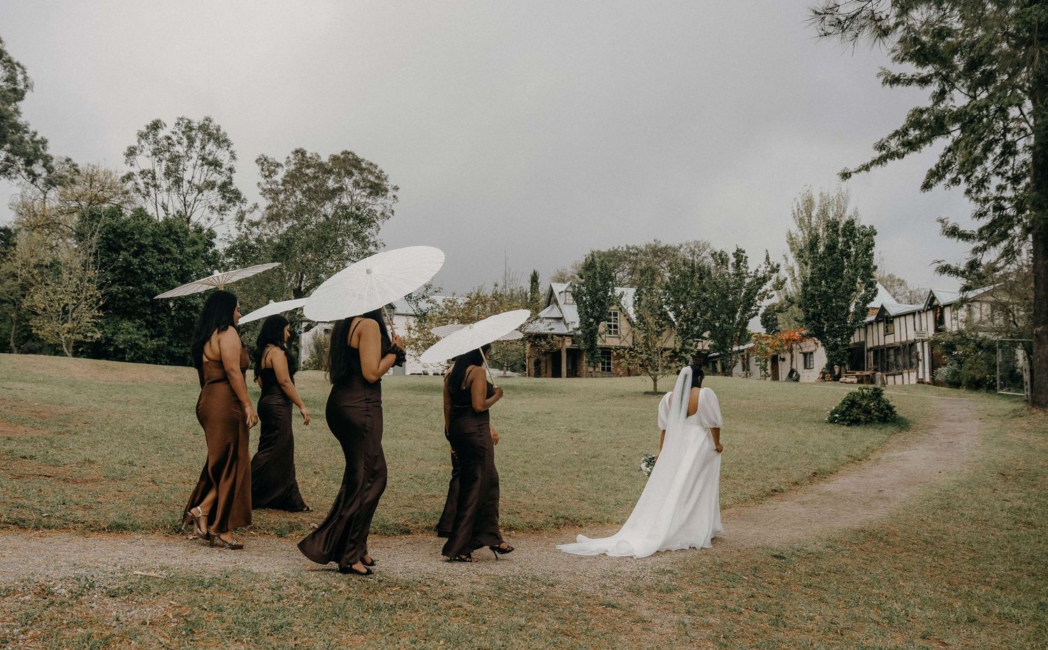Bridesmaids in dark dresses lift their bouquets over the bride walking outdoors on an overcast day.