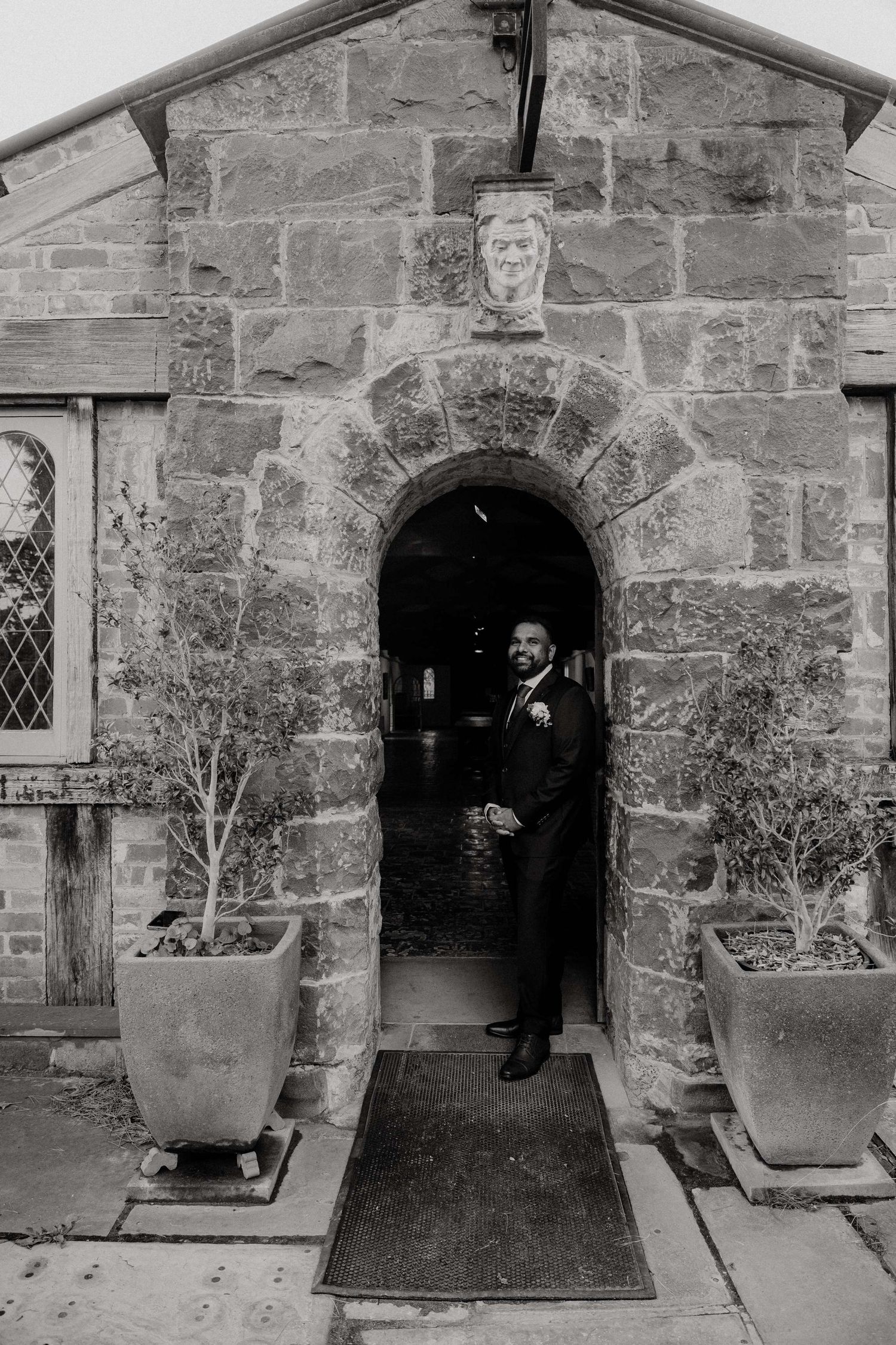 A person in formal attire stands in a stone archway entrance of a historic building with decorative planters on either side.