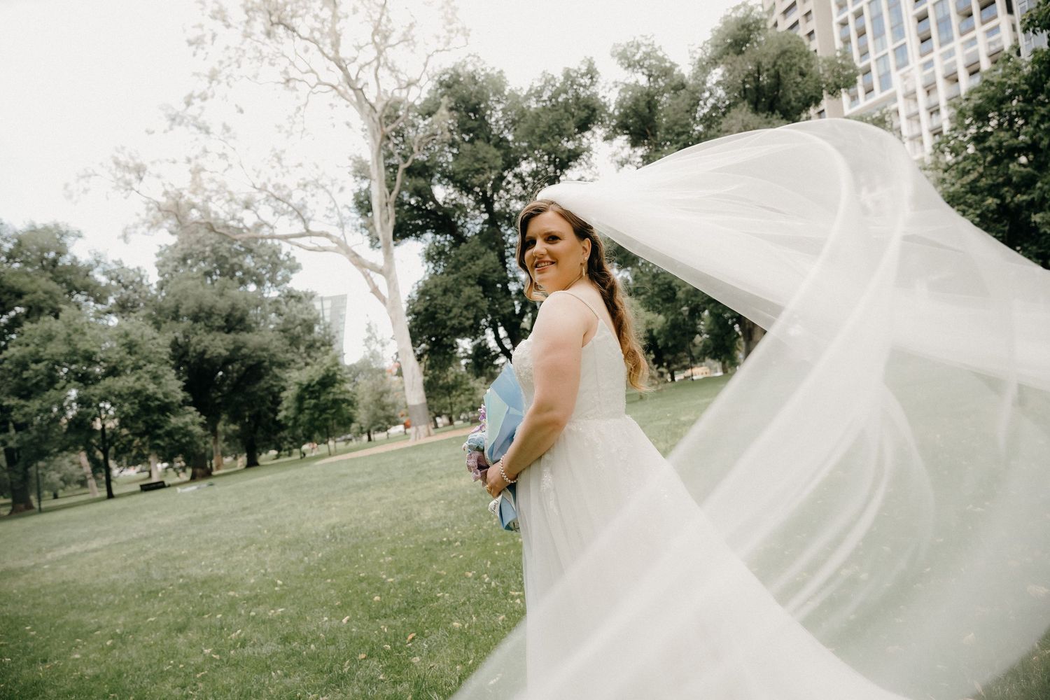 A flowing white wedding veil billows gracefully in the wind against a lush green park background.