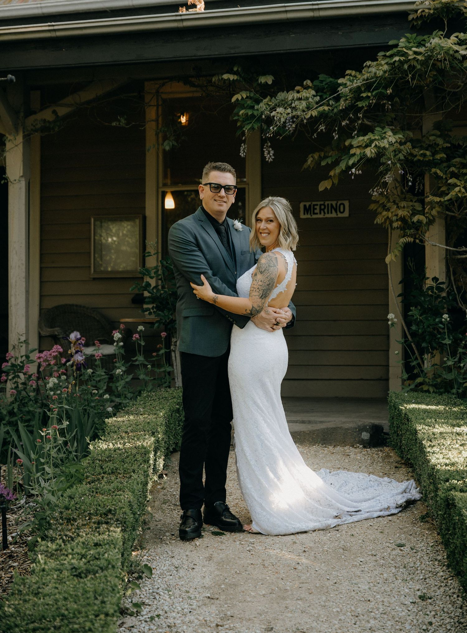 A couple in wedding attire embraces on a garden path in front of a rustic dark green house with blooming flowers.