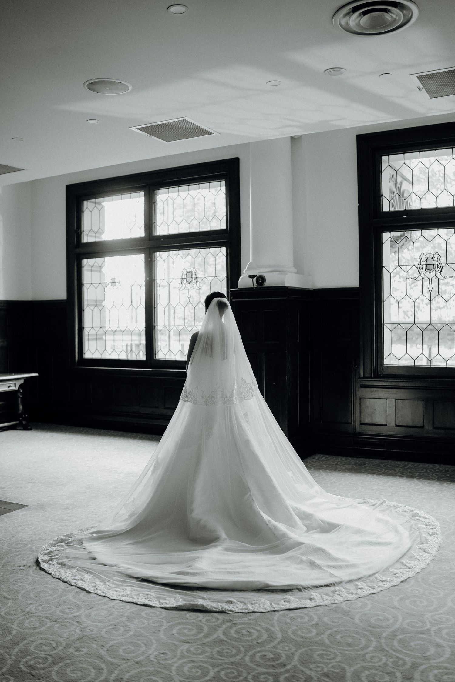 Black and white photo of a bride in a flowing white gown and long veil standing by leaded glass windows.