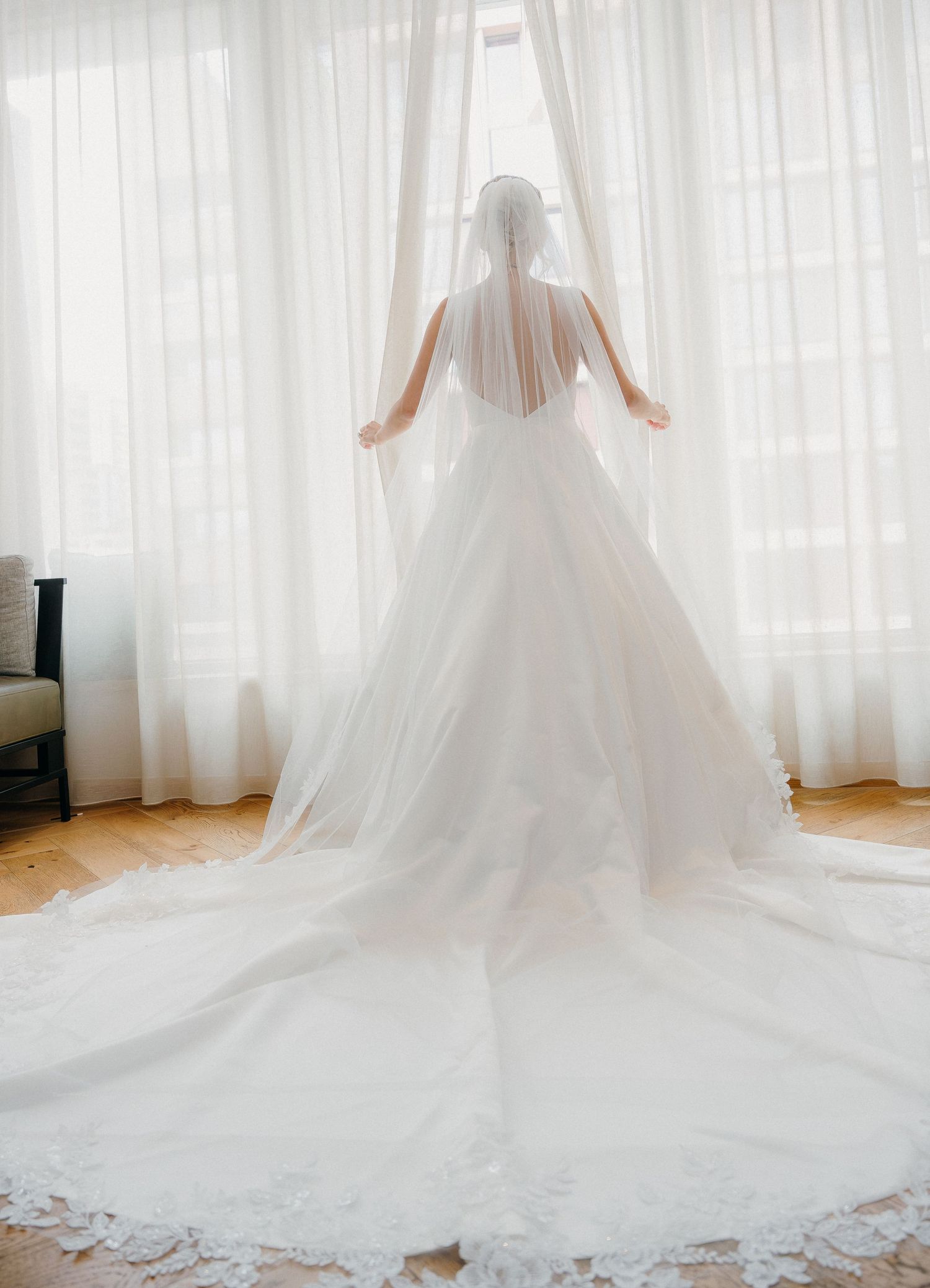 A stunning white wedding dress with a long cathedral train spreads across a sunlit room with sheer curtains.