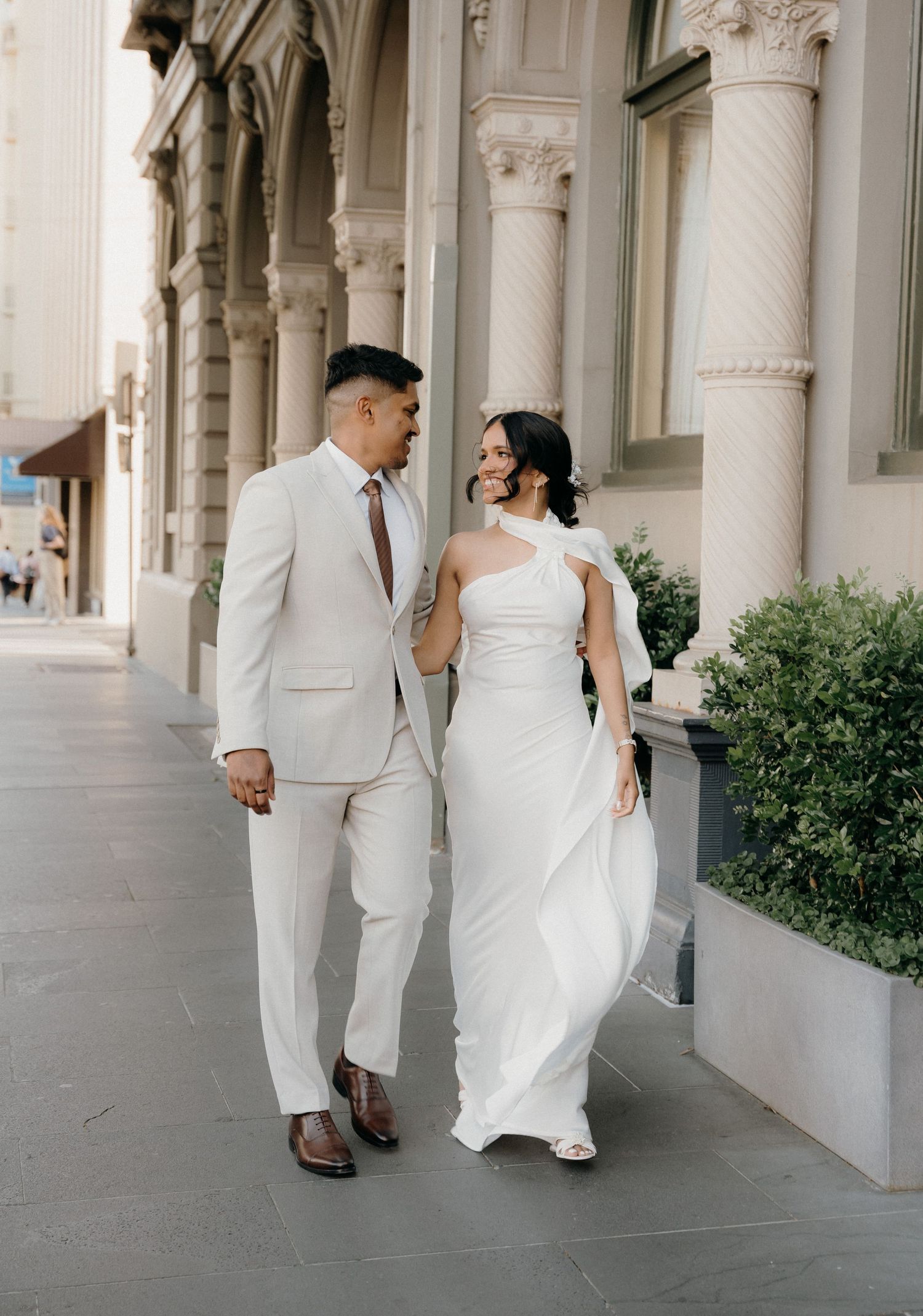 A wedding party dressed in elegant white attire walks through a classic colonnade with ornate pillars and greenery.