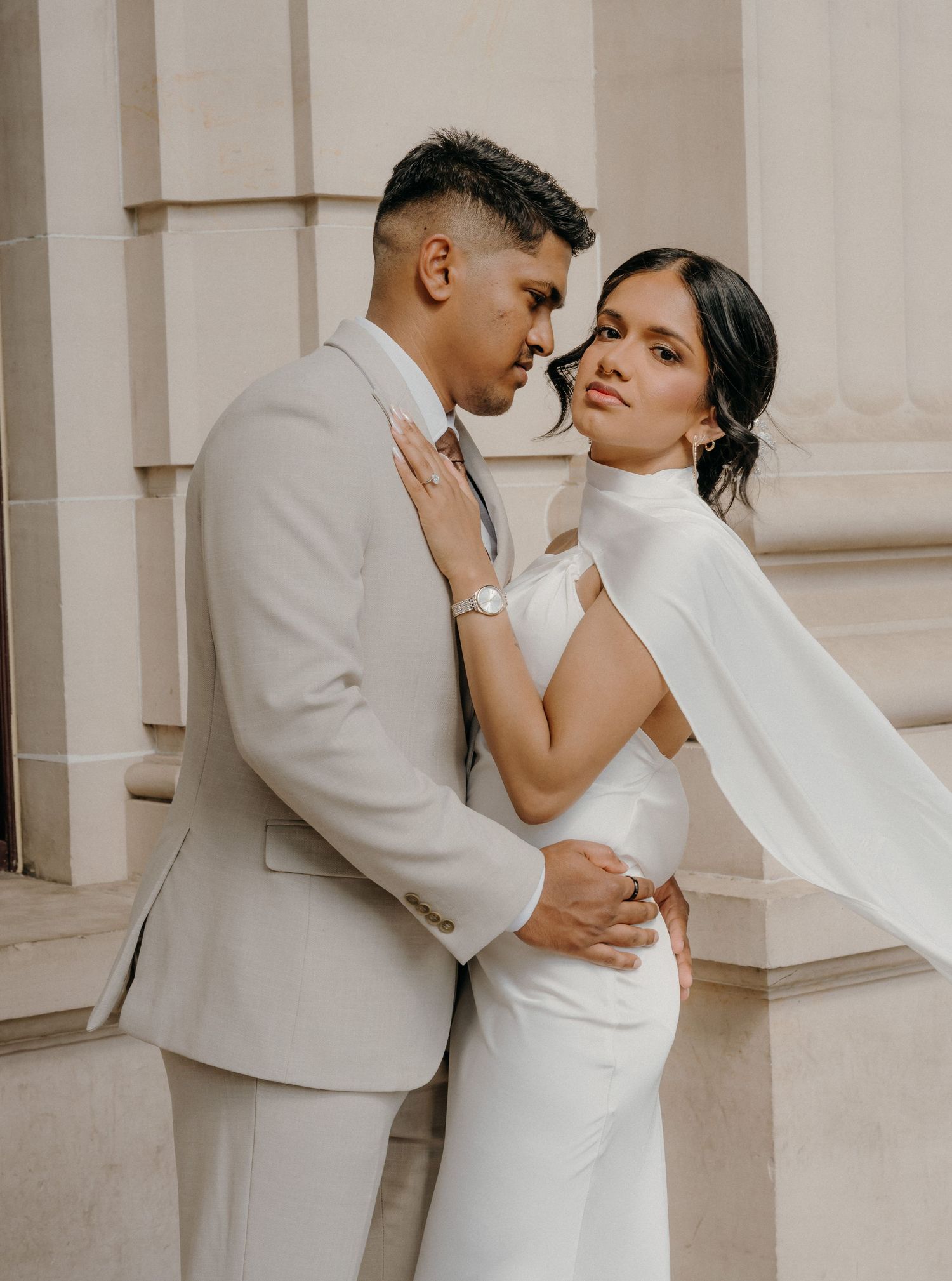 A romantic wedding couple embraces against a light colored wall in matching white outfits.